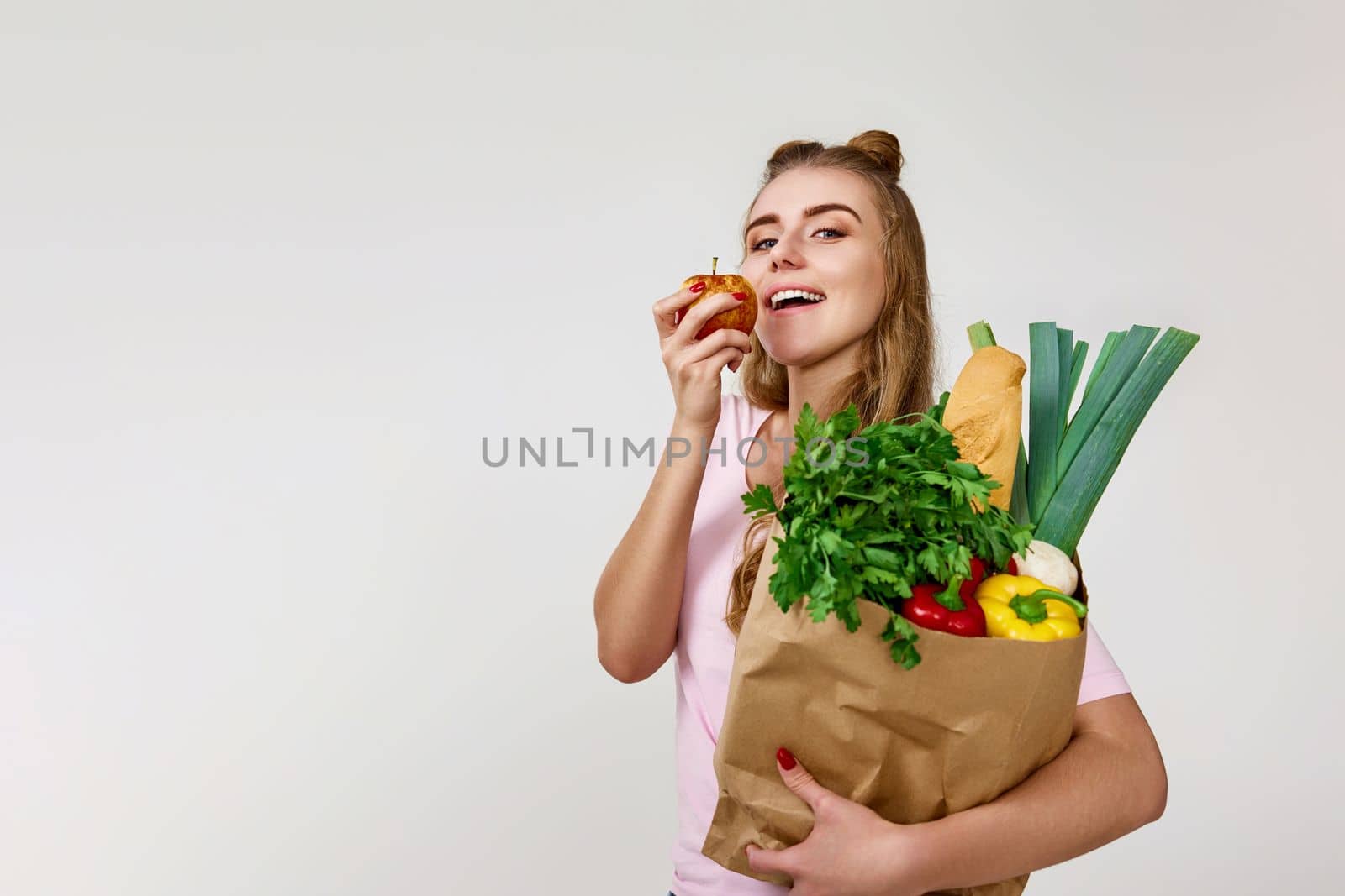 caucasian young woman in pink t-shirt with paper bag with vegetables biting apple. copy space. healthy food for diet