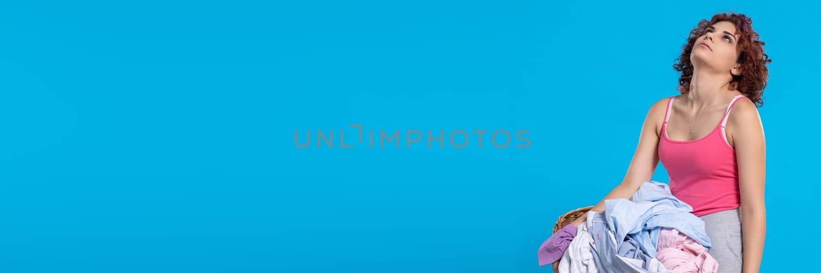 A very tired woman , carries a heavy wicker basket with washed clothes. Overworked woman with dark and curly hair. Monotonous housework. . Panoramic view of girl with empty blue background.