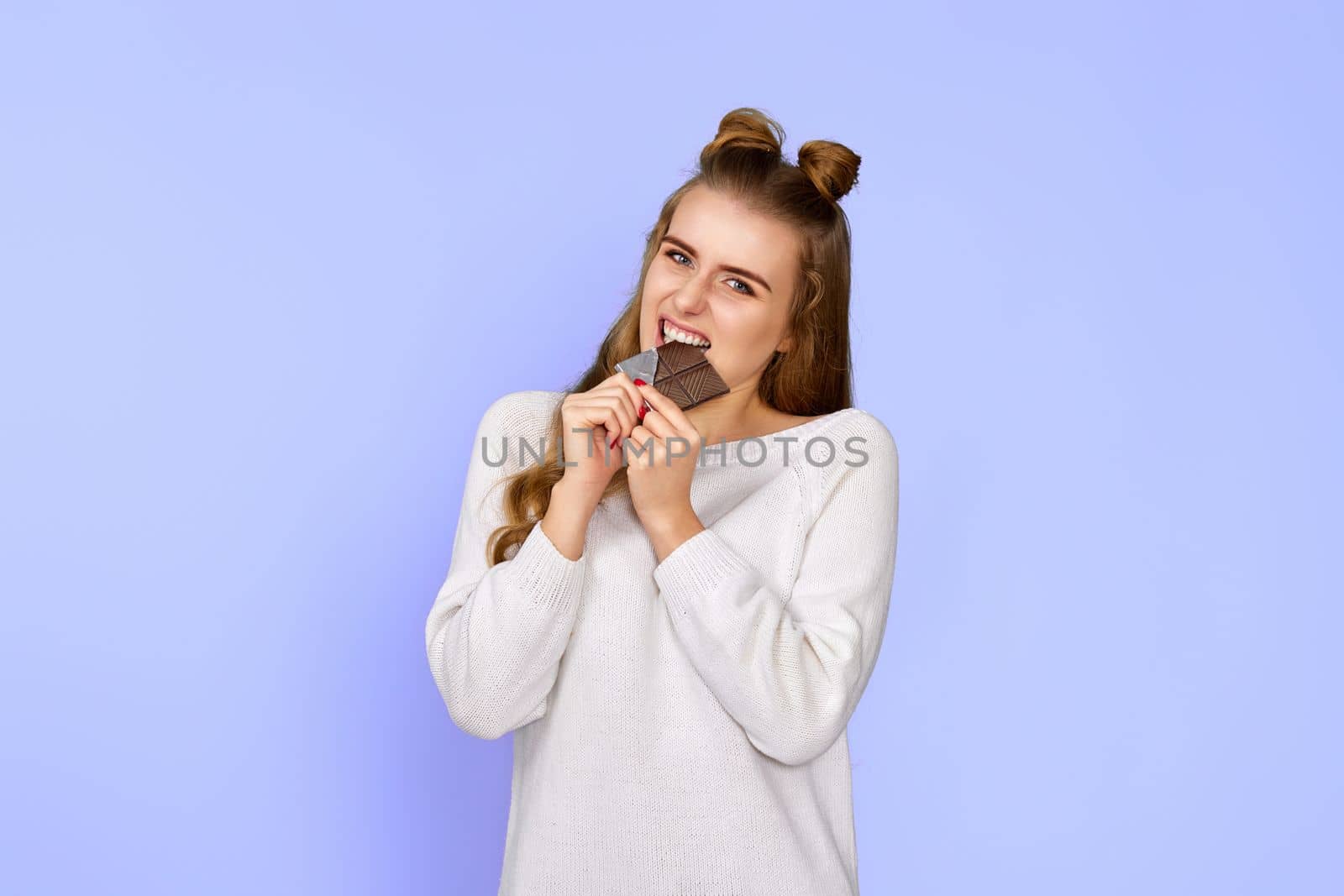 young happy woman in sweater biting sweet chocolate bar and looking at camera isolated on pastel violet background