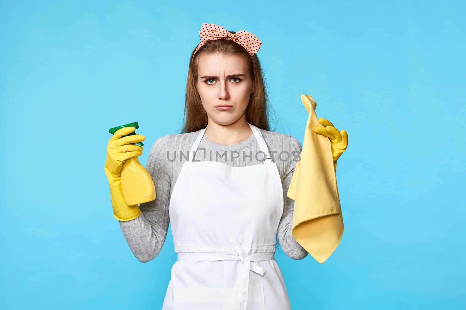 skeptic and nervous, frowning woman in gloves and cleaner apron with cleaning rag and detergent sprayer on blue background.