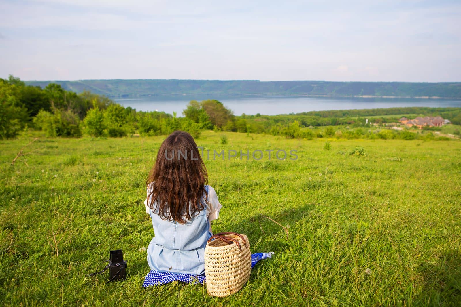 A very beautiful view of the lake, a girl is sitting on a blanket.. A straw picnic basket is standing nearby on the green grass along with an old camera. Recreation in the fresh air. by sfinks