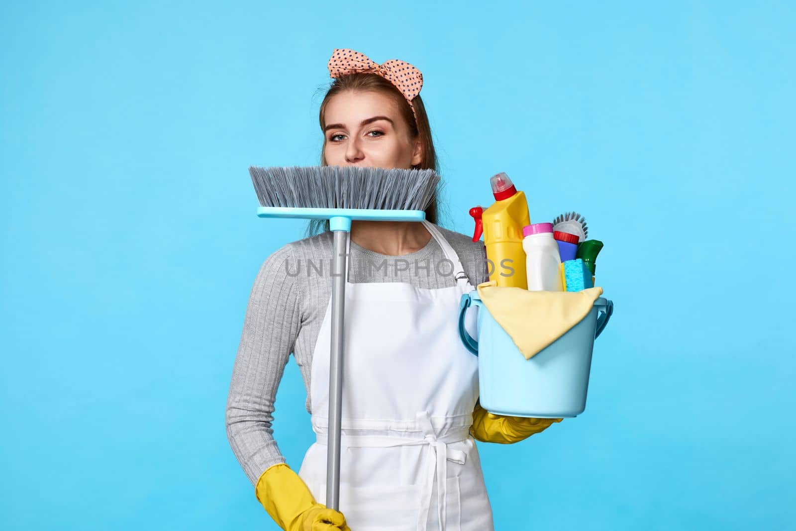 smiling woman in yellow rubber gloves and cleaner apron holding bucket of detergents and broom on blue background.