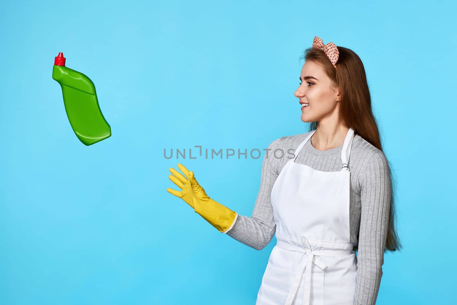 caucasian woman in yellow rubber gloves catches flying bottle of detergent on blue background.