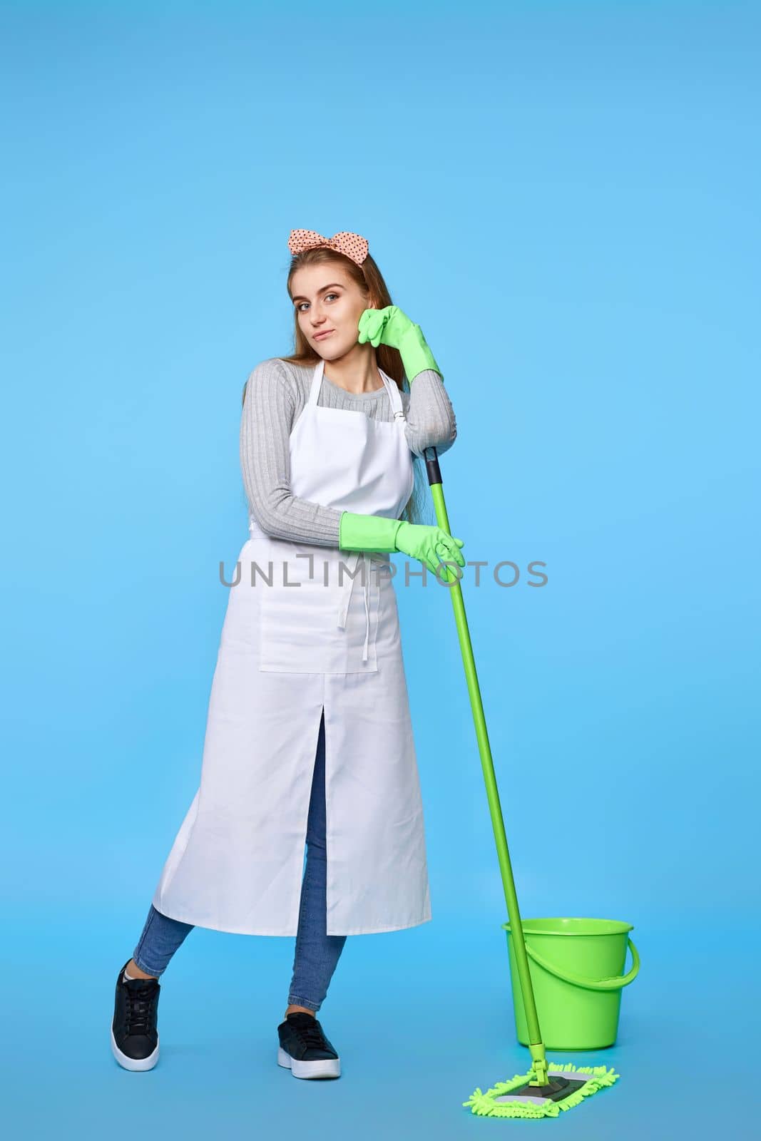 pretty woman in green rubber gloves and cleaner apron washing floor with mop on blue background. Full length