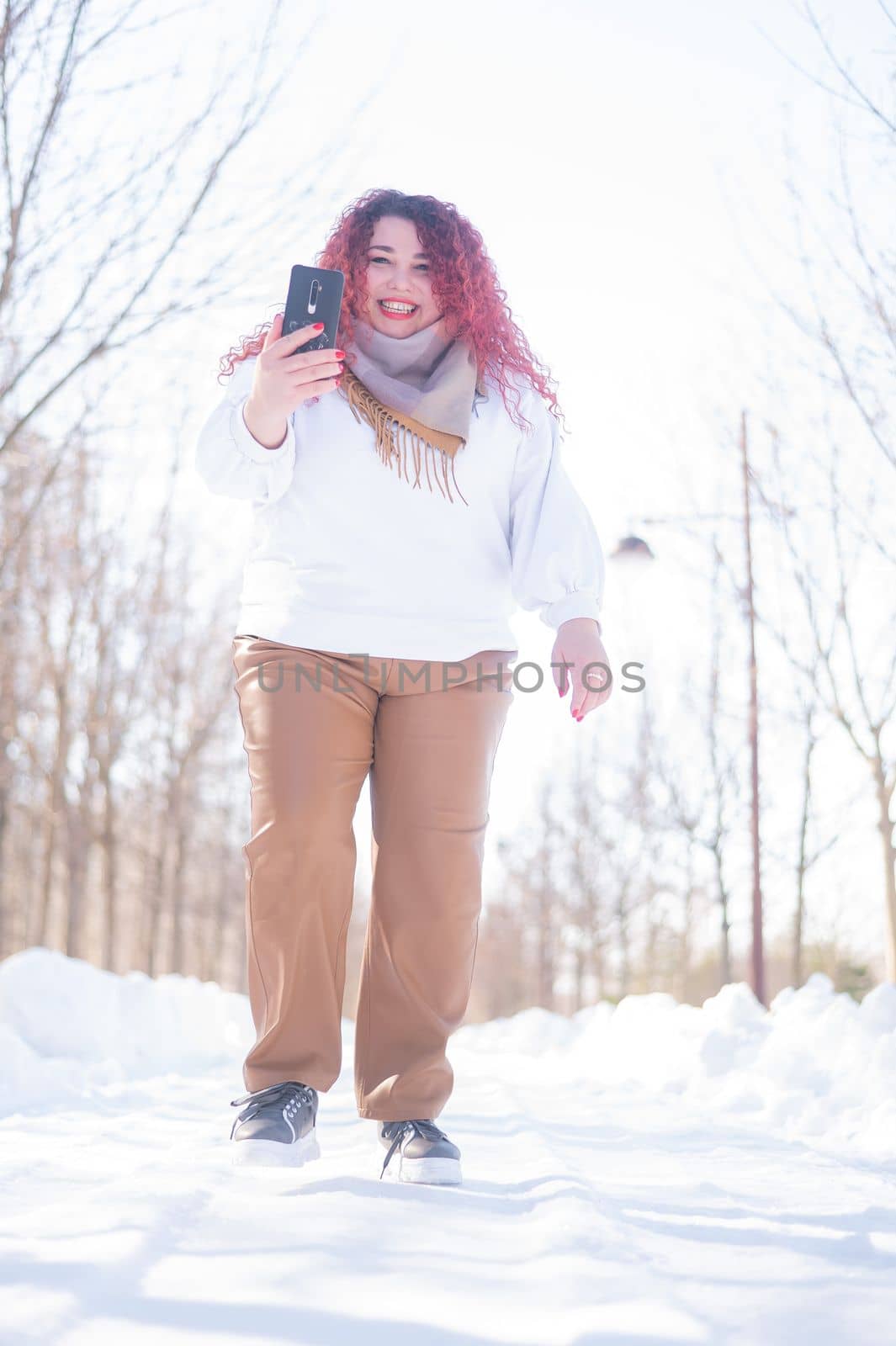 Portrait of a smiling plump red-haired woman taking a photo on a smartphone on a walk in winter. by mrwed54