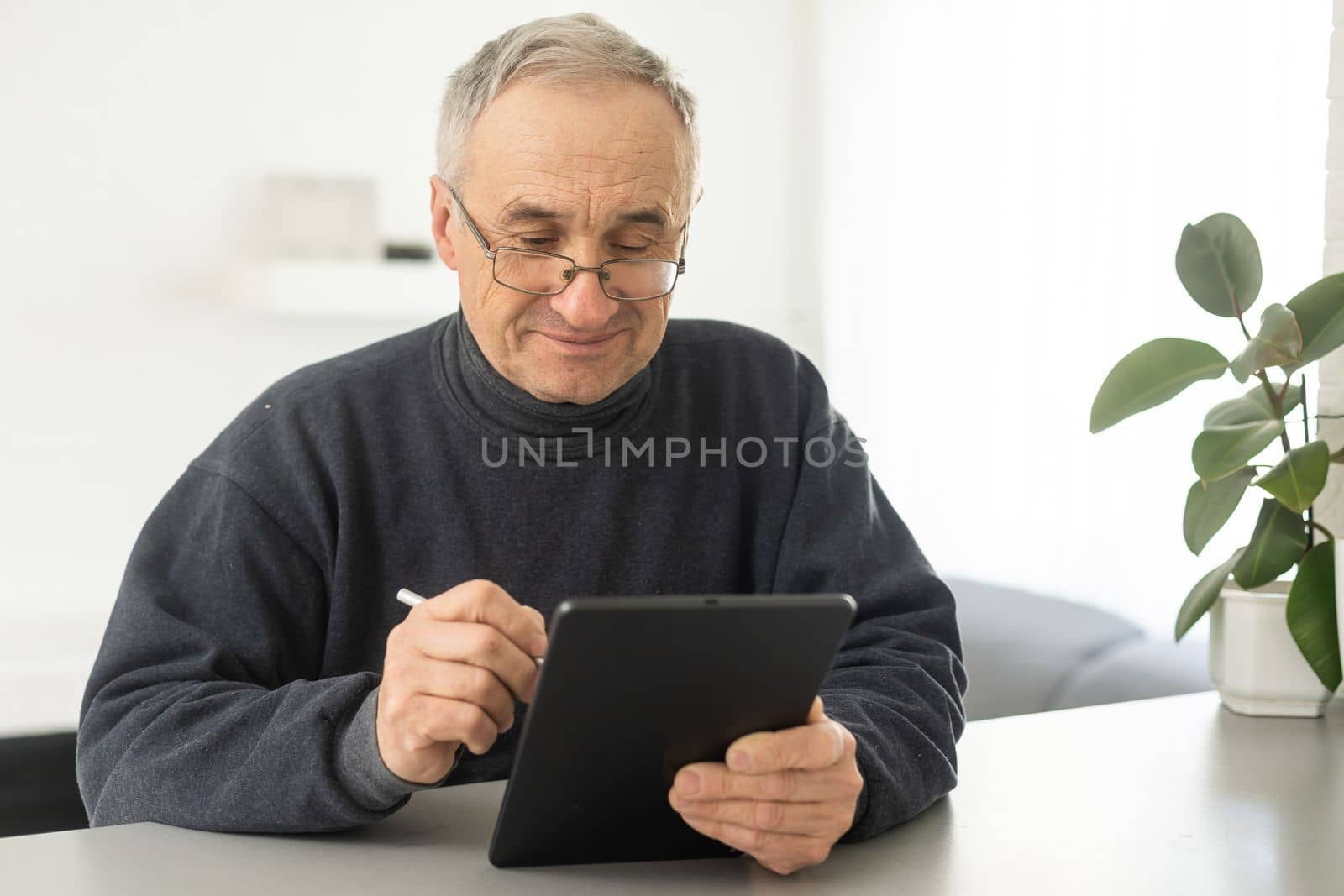 Senior man reading news on digital tablet. Serious mature male using portable computer at home, sitting at table, copy space. by Andelov13