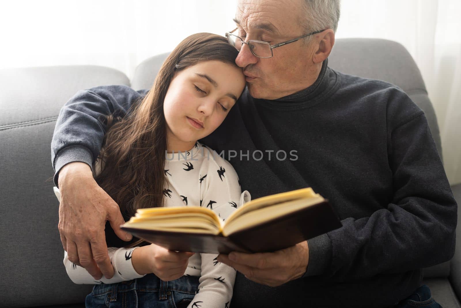 Photo of aged old grandpa little pretty granddaughter sit sofa hugging stay house quarantine safety read interesting book fairy tale modern interior living room indoors. by Andelov13