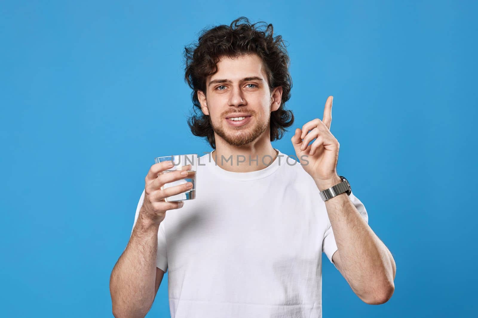 Young man in white t-shirt with drinking glass of water pointing up on blue background