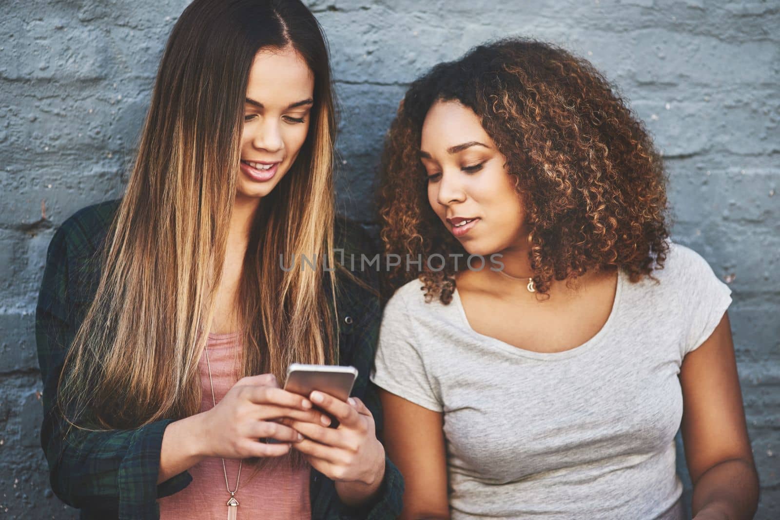 How todays youth keep up with the current trends. two young women standing outdoors and using a mobile phone against a gray wall
