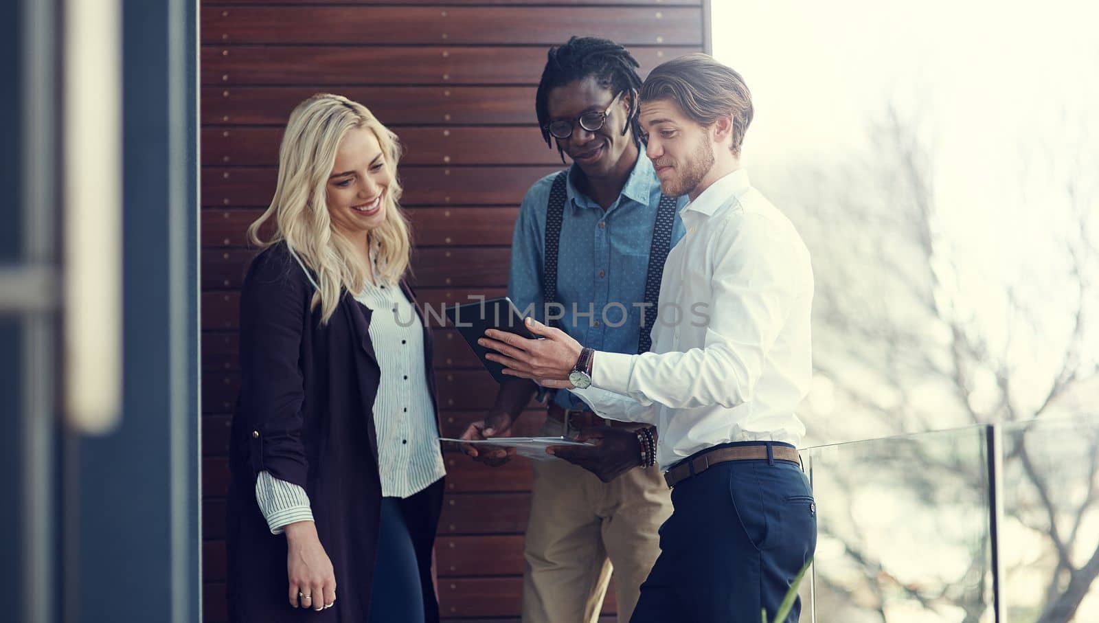 Getting some advice from trusted colleagues. three young businesspeople using a tablet while standing outside on the office balcony. by YuriArcurs