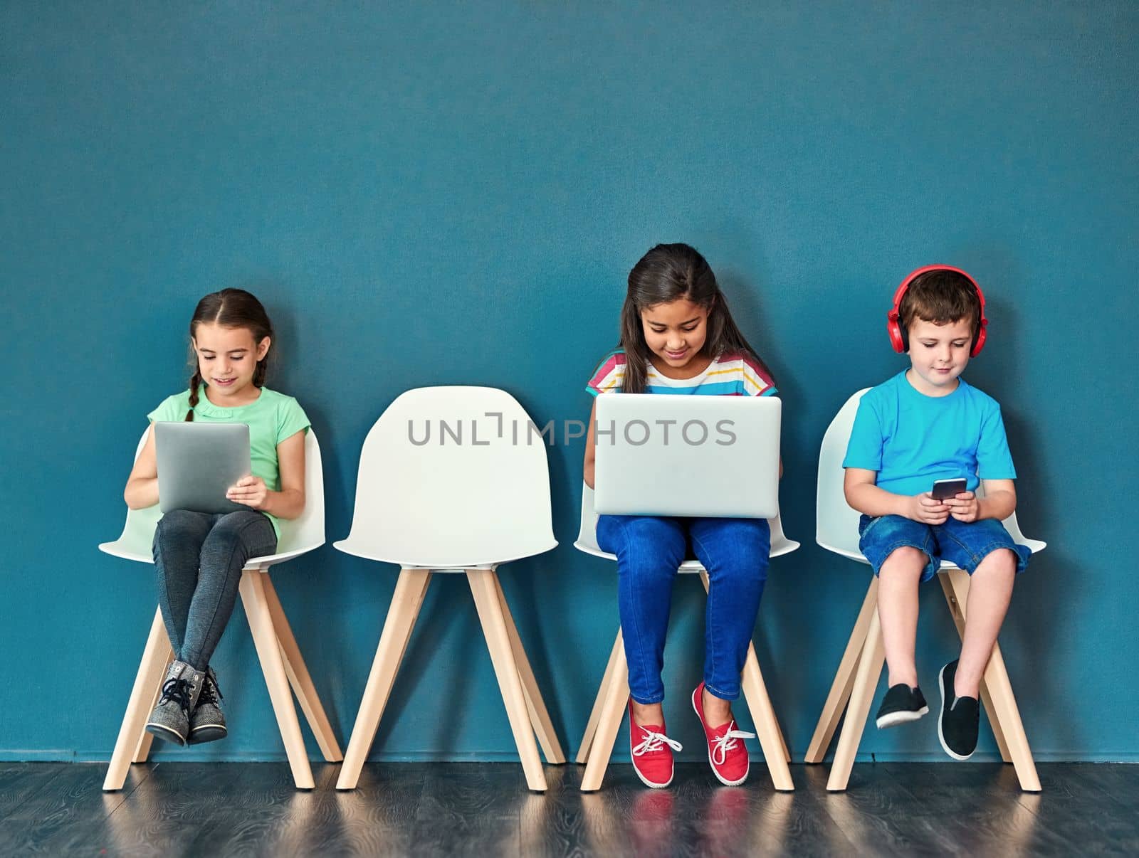 Growing up in a wireless world. Studio shot of kids sitting on chairs and using wireless technology against a blue background
