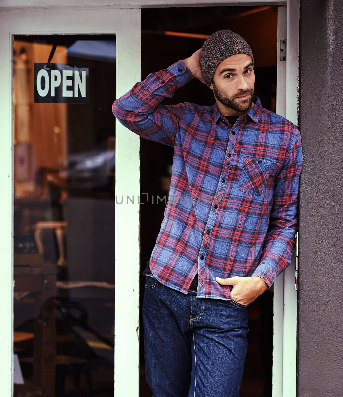 Yes we are open for business. a handsome young man posing outside a cafe with his one hand in his pocket and the other behind his head. by YuriArcurs