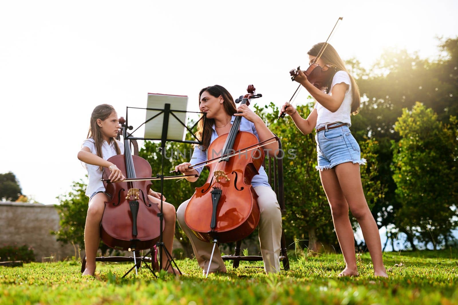 What a great way to spend quality time with the kids. a cheerful young mother and her two daughters playing classical string instruments together while standing in the backyard of their home