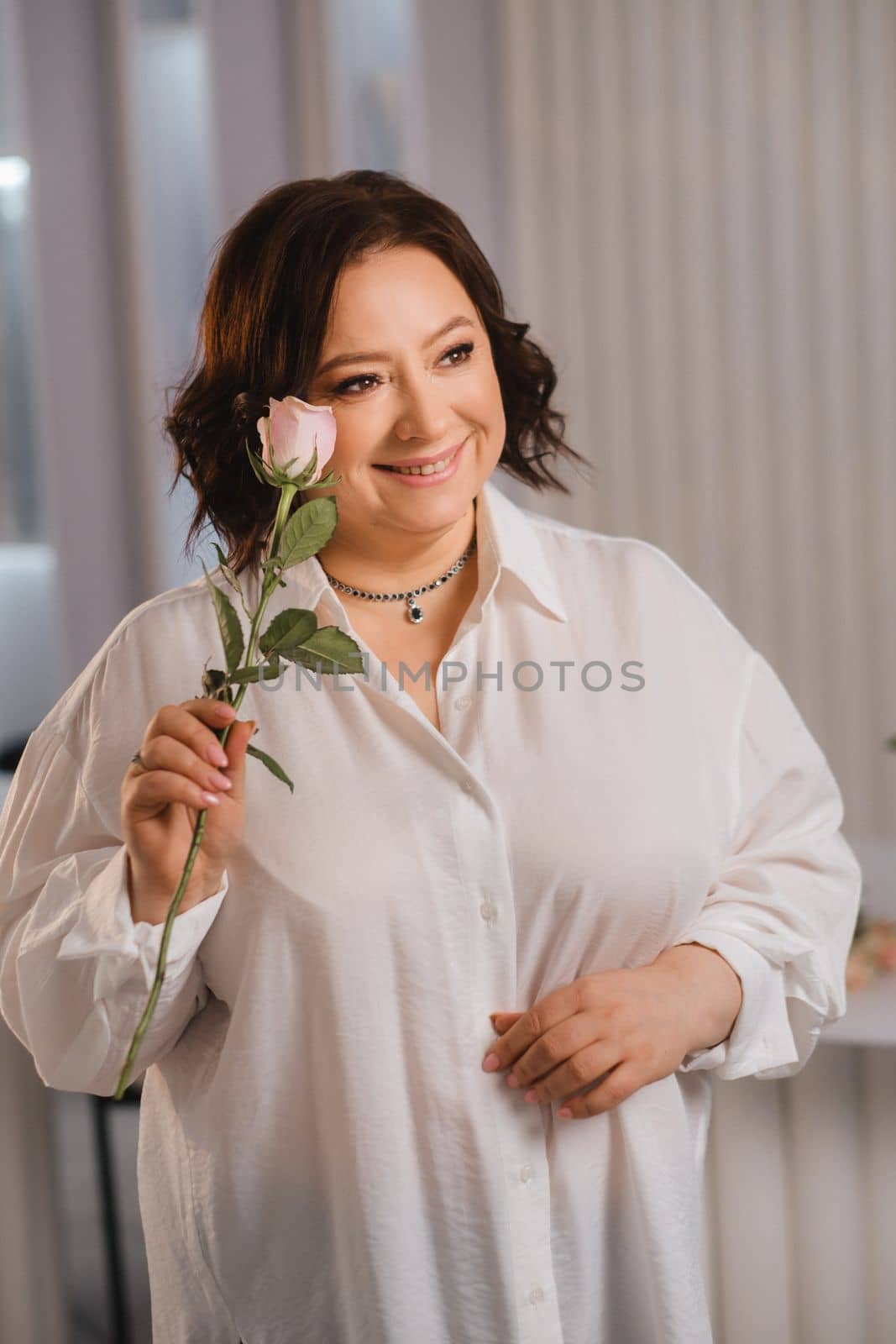 An adult woman in a white shirt holds one rose in her hands while standing in the interior. Pink roses.