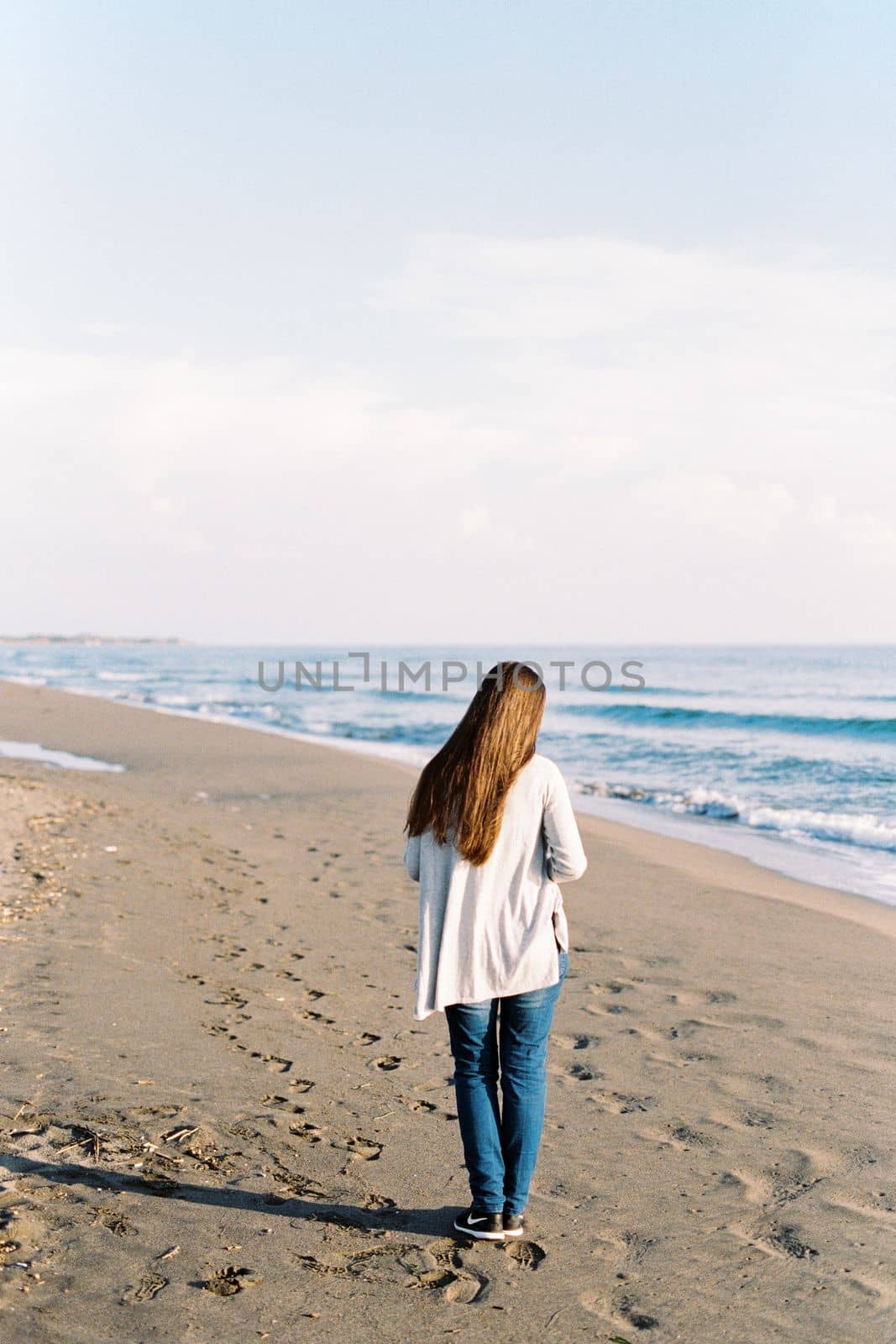 Young woman in a long sweater walks along the sandy beach. Back view. High quality photo