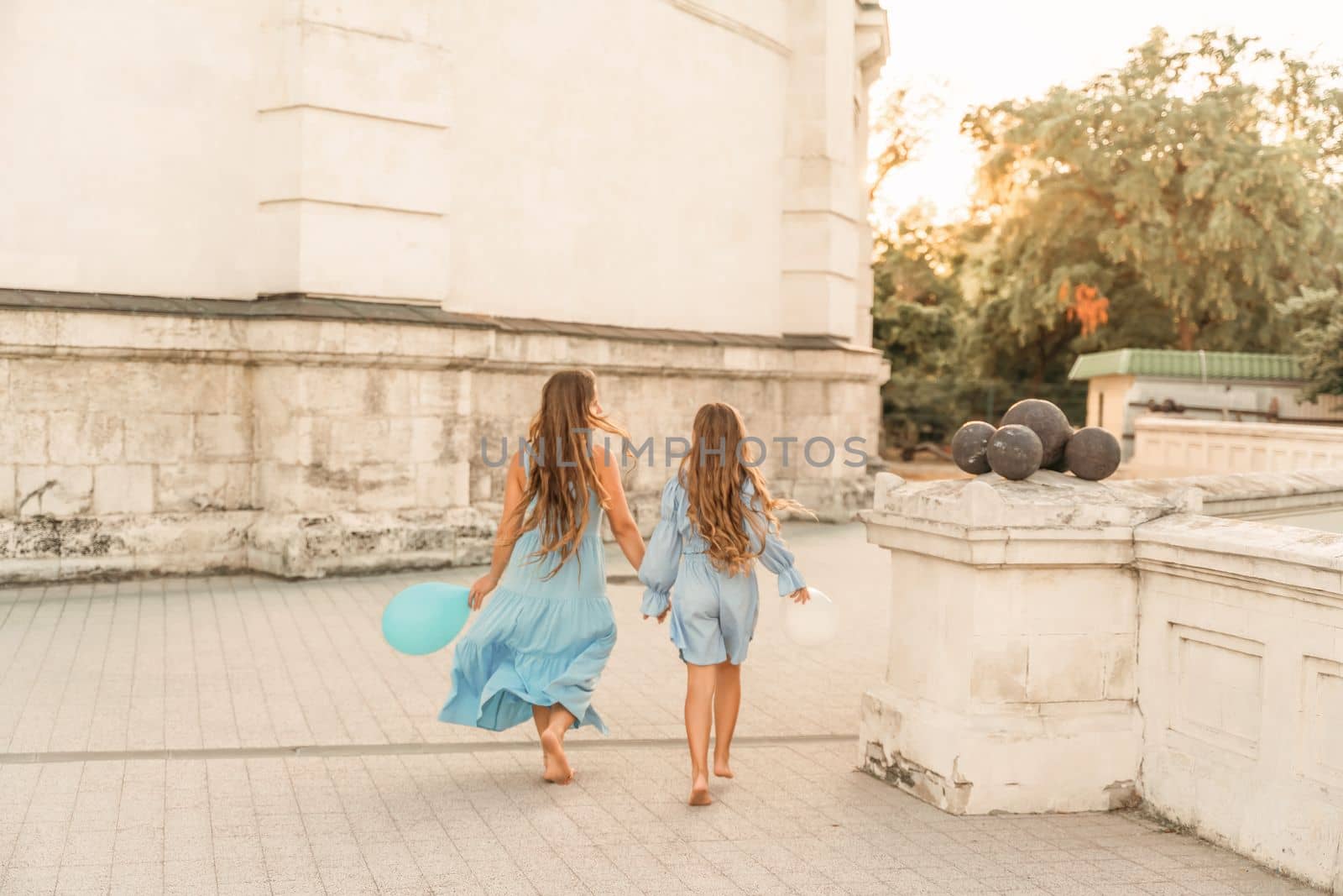 Daughter mother run holding hands. In blue dresses with flowing long hair, they hold balloons in their hands against the backdrop of a sunset and a white building. by Matiunina