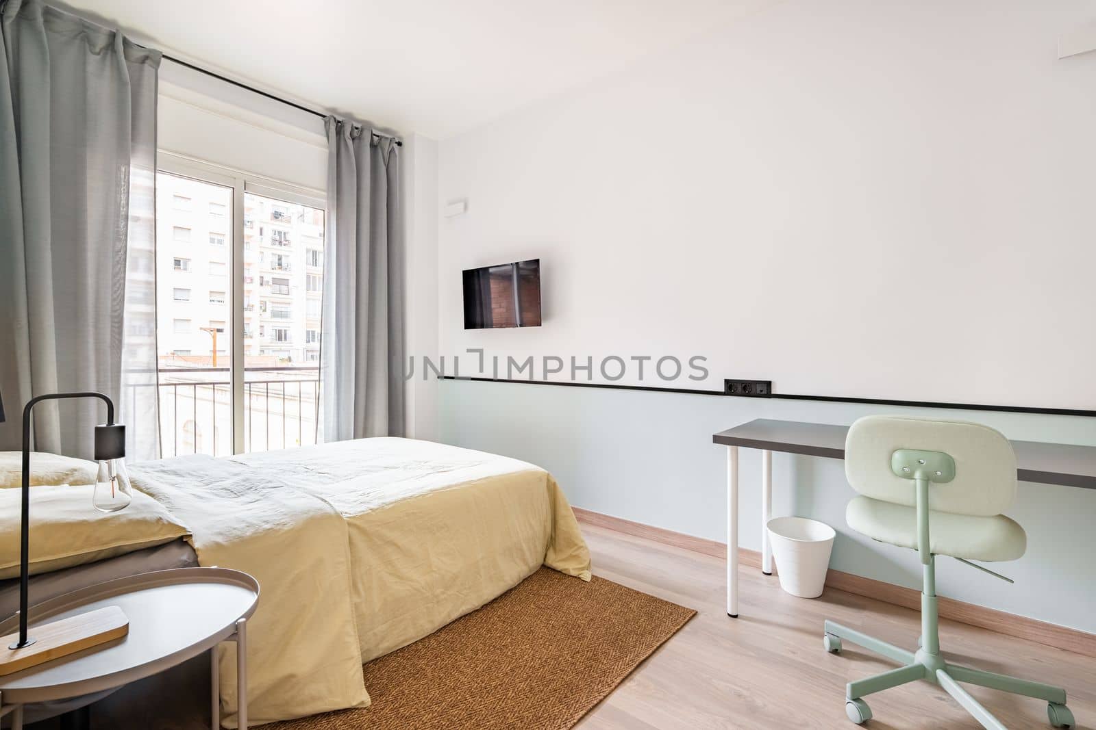 Hotel room with bed for two persons with beige linen. Access to balcony through glass sliding doors. Table against wall with chair for online work. On wall is TV for pleasant evenings watching movie. by apavlin