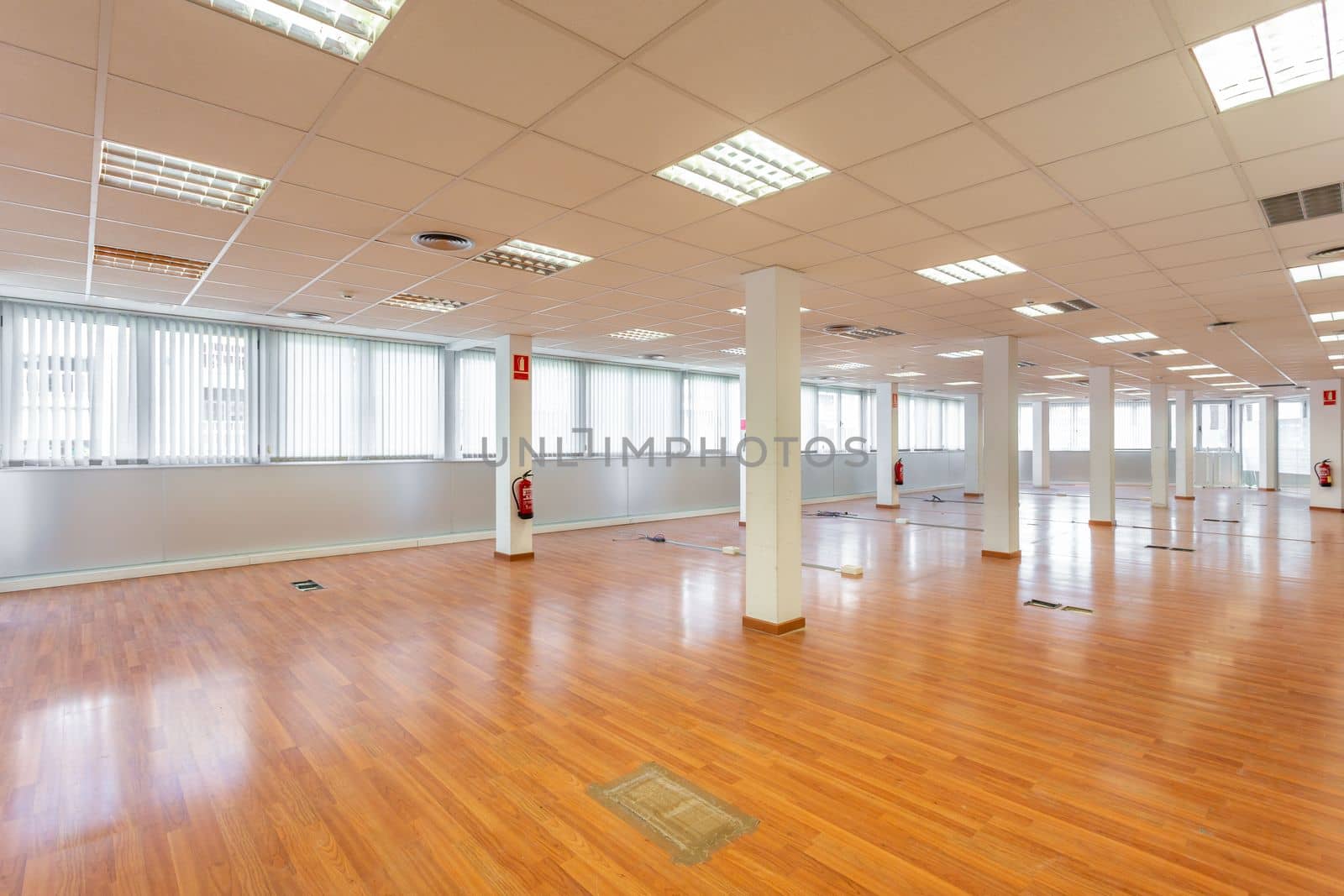Large empty space with ceiling tiles, fluorescent lights, light brown laminate flooring and white painted columns. Large office space left from a company