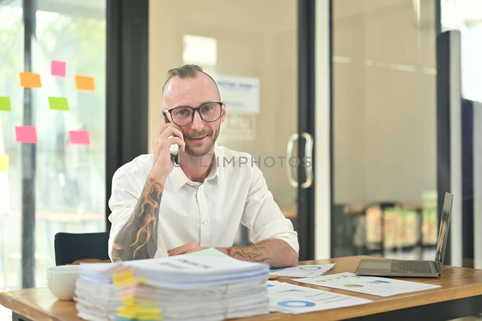 Positive caucasian start up businessman talking on mobile phone with business partners while sitting at workplace by prathanchorruangsak