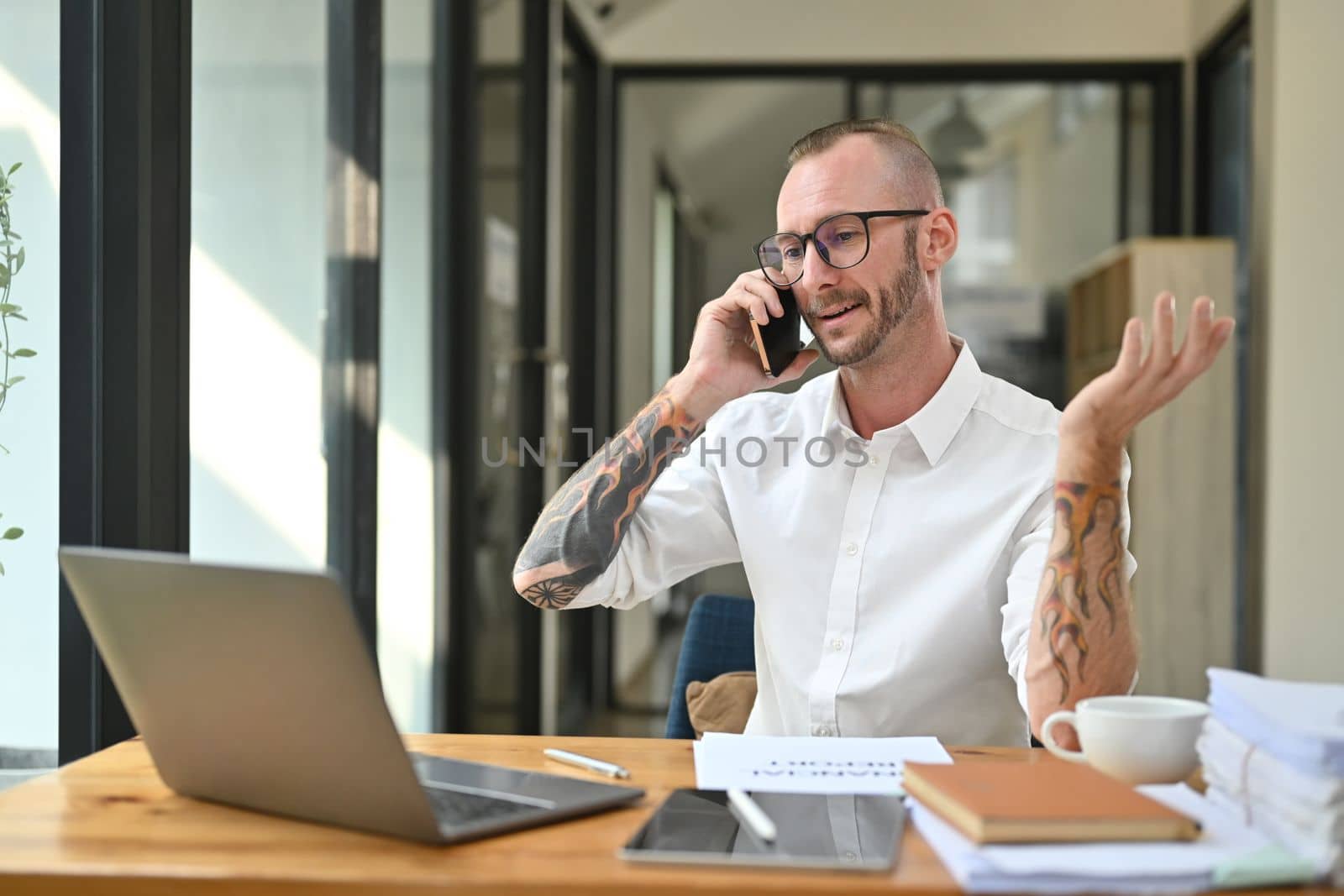 Friendly caucasian male manager wearing glasses talking on phone, sitting at desk with laptop.