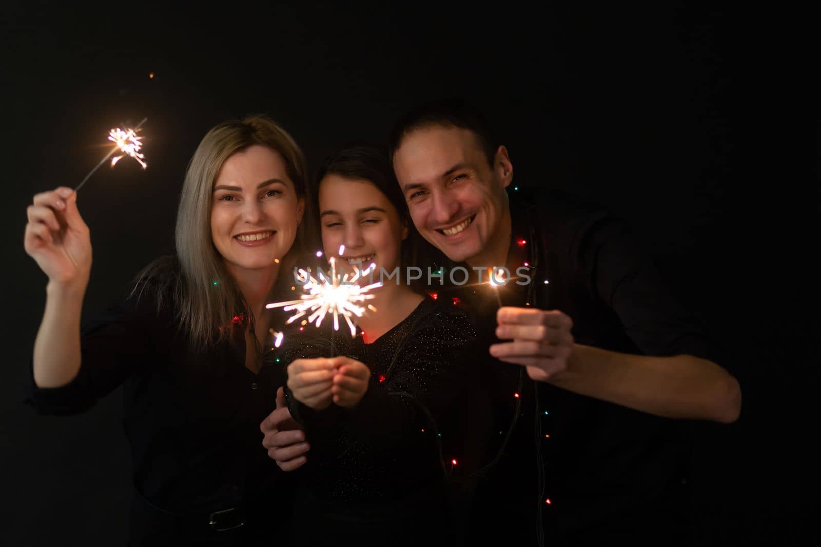 Happy family in black - woman, man and little girl, with a sparkler in hand the New Year's Christmas at home. The girl at the father on hands. New year 2023