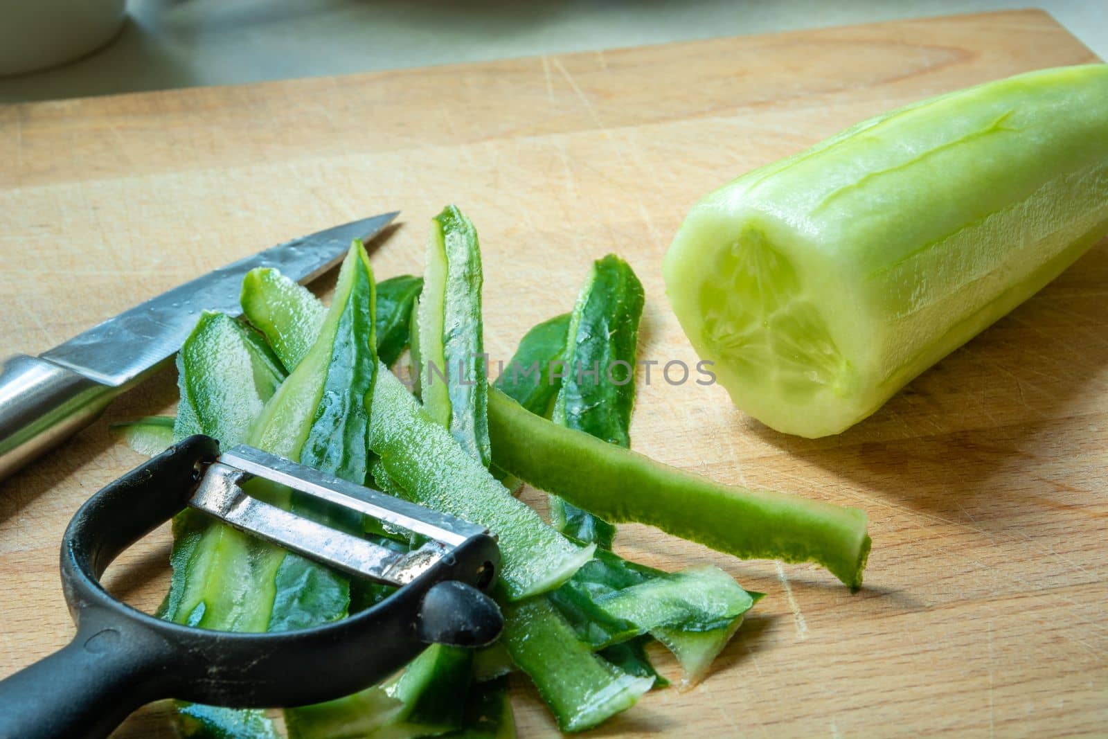 Vegetable peeler and knife lying next to peeled green cucumber
