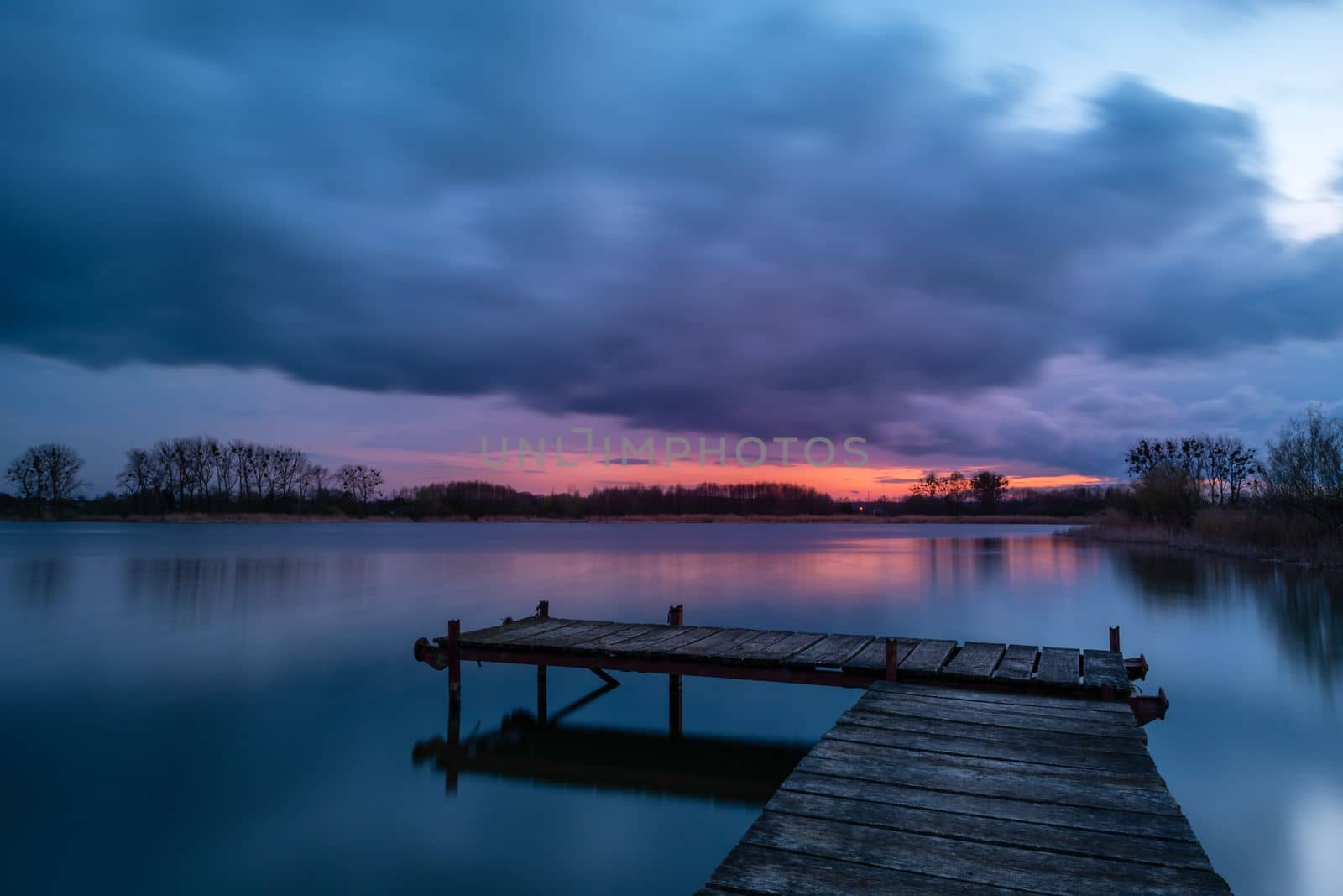 View from a wooden pier on a calm lake during sunset with dark clouds by darekb22