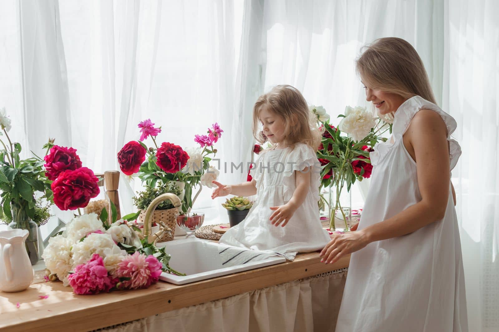 A little blonde girl with her mom on a kitchen countertop decorated with peonies. The concept of the relationship between mother and daughter. Spring atmosphere.