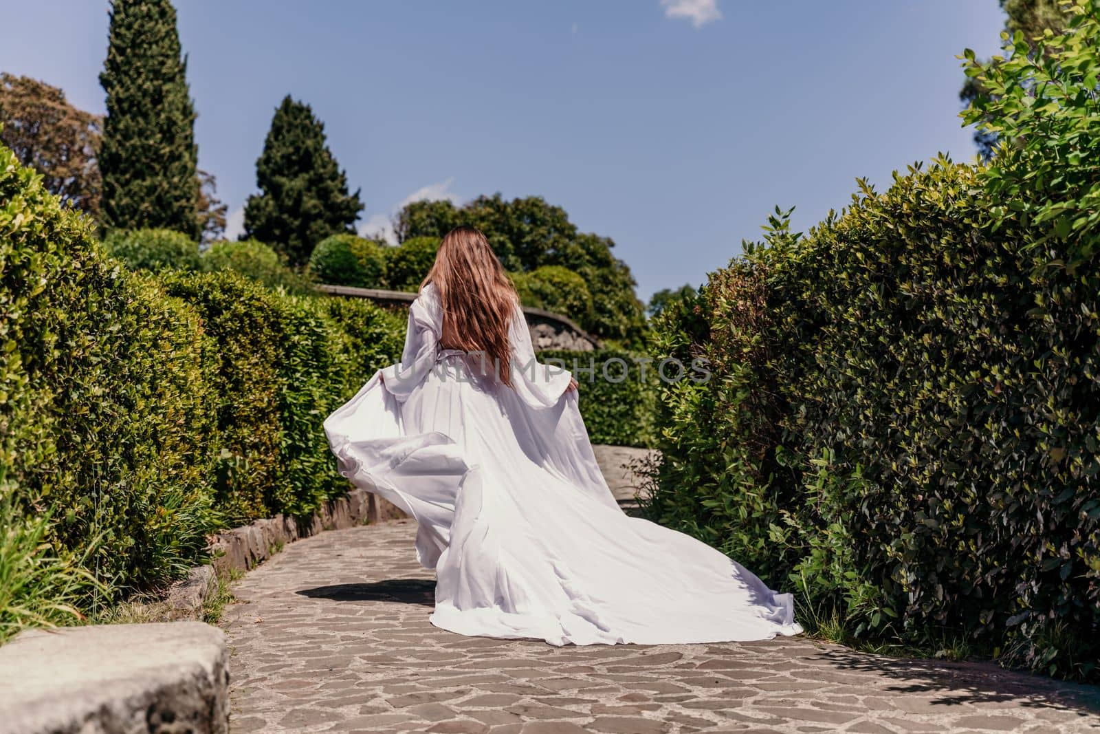 Brunette runs white dress park. A beautiful woman with long brown hair and a long white dress runs along the path along the beautiful bushes in the park, rear view by Matiunina