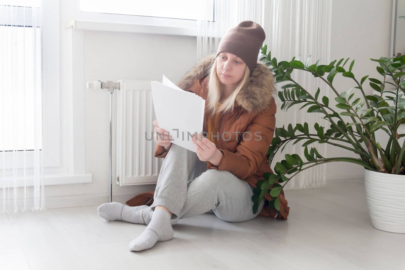 A woman warmly dressed in a brown jacket and hat is sitting near the heater with bills in her hands. The concept of a crisis and a large set for utilities