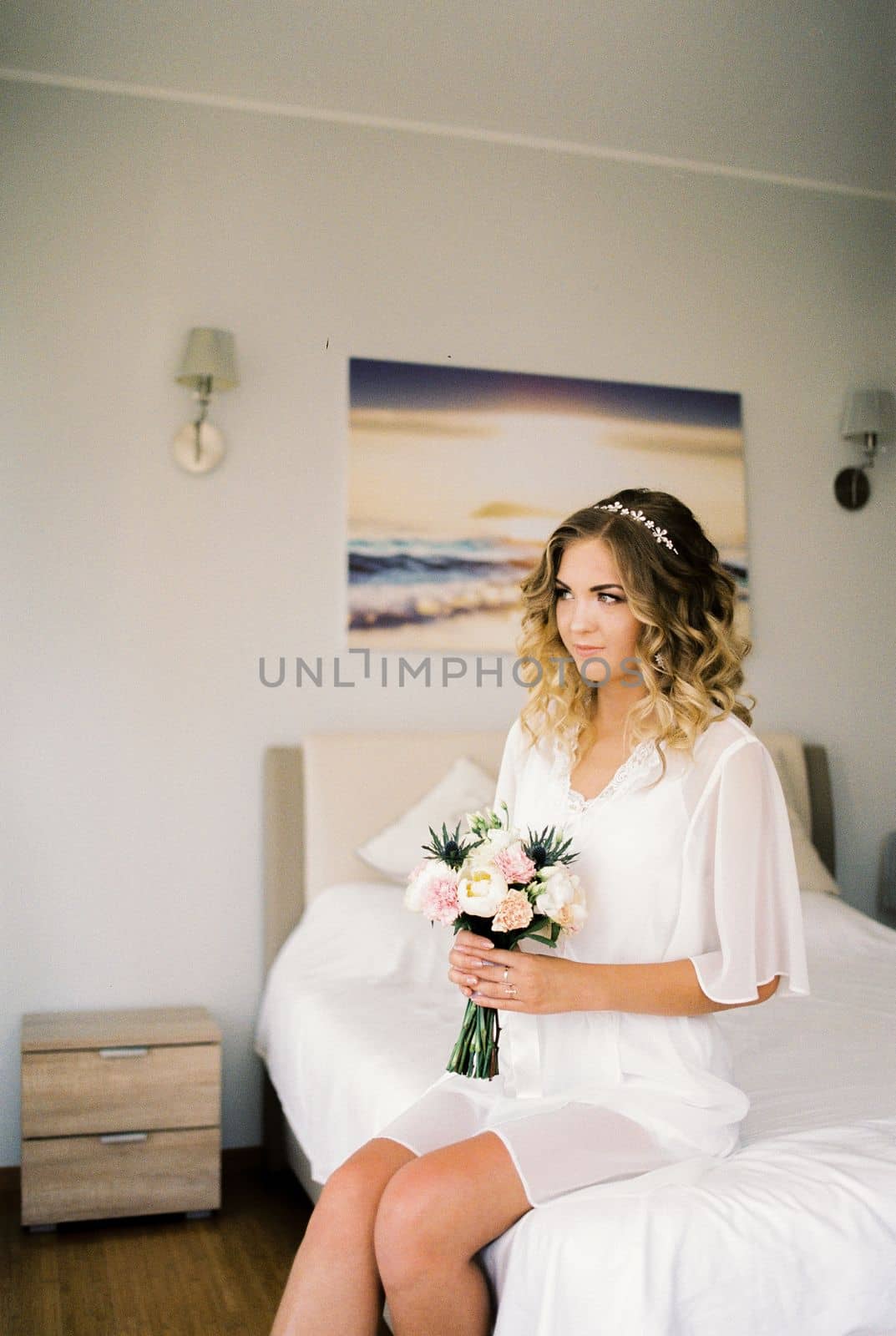 Bride in a white bathrobe sits with a bouquet of flowers on the bed in the room. High quality photo