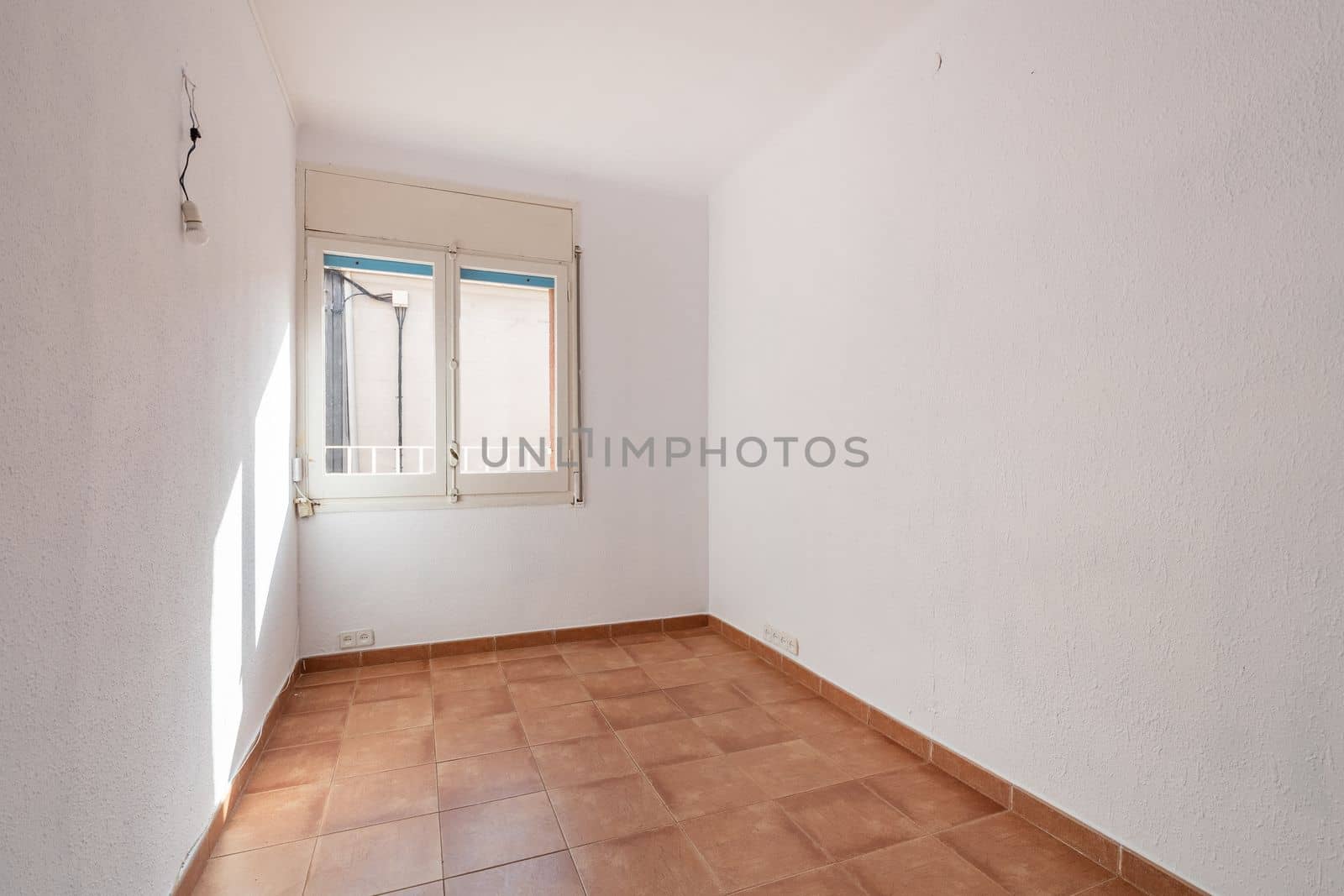 An empty spacious room with old square worn caramel colored floor tiles and window with natural sun light, white faded walls and ceiling. The wires for the lamp stick out in the wall. by apavlin