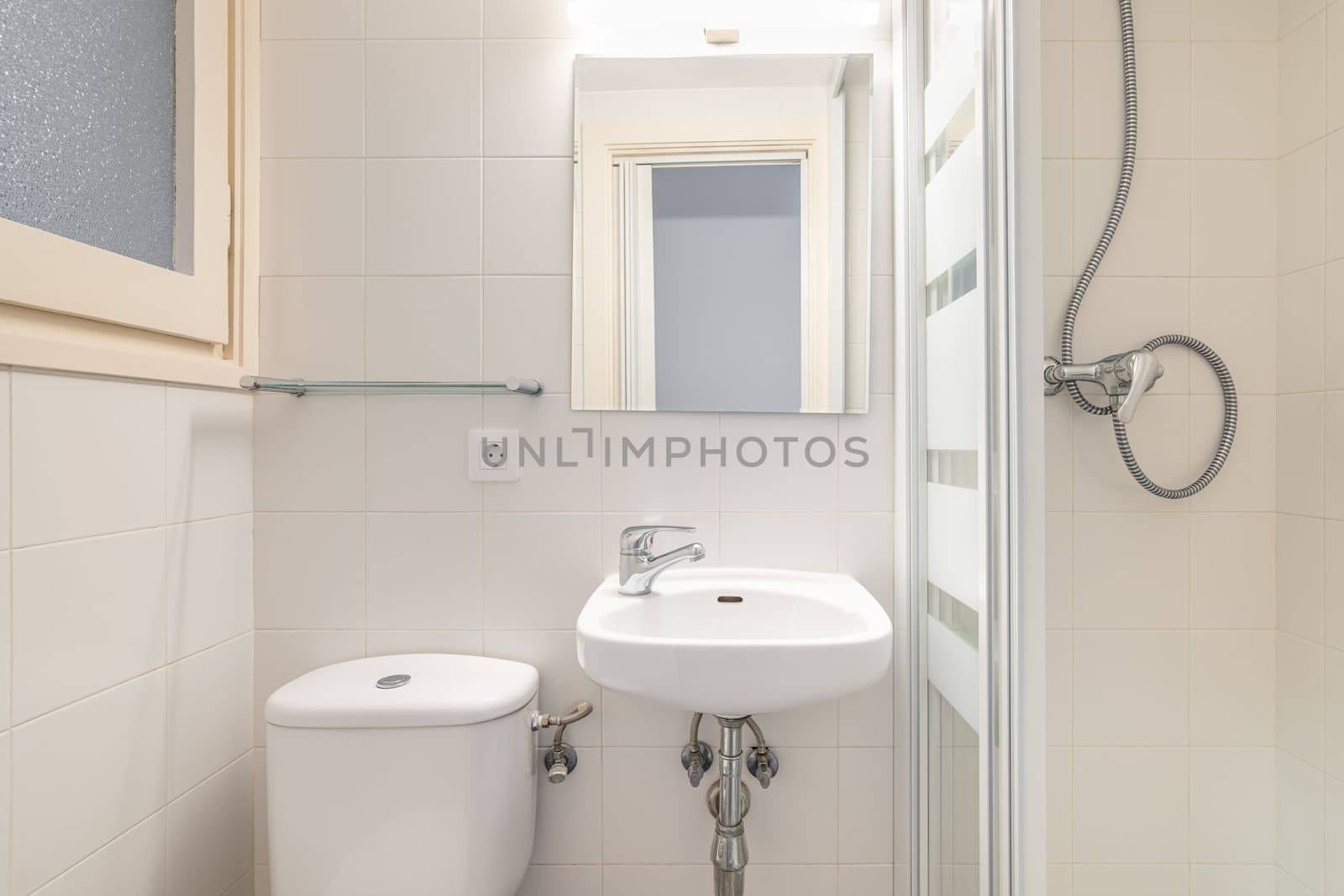 Bathroom with light beige walls. In corner is a shower with glass sliding doors. Vanity sink with white furniture. Mirror is illuminated by bright lamps with a reflection of the front door