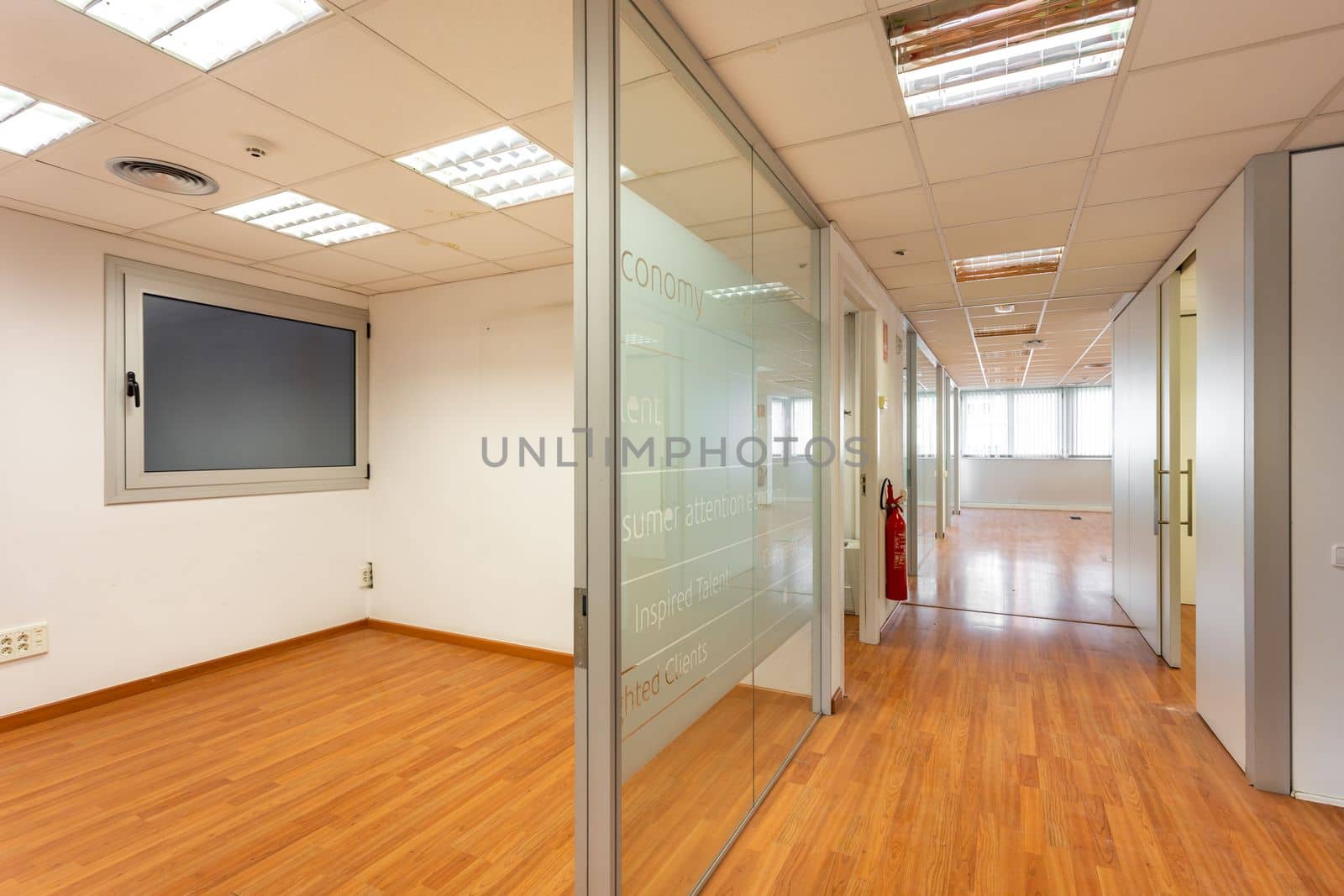 Glass partition enclosing empty office room from corridor with window at end. Outdated design of office corridor and room that needs to be renovated. Burnt out fluorescent lamps in empty office space. by apavlin