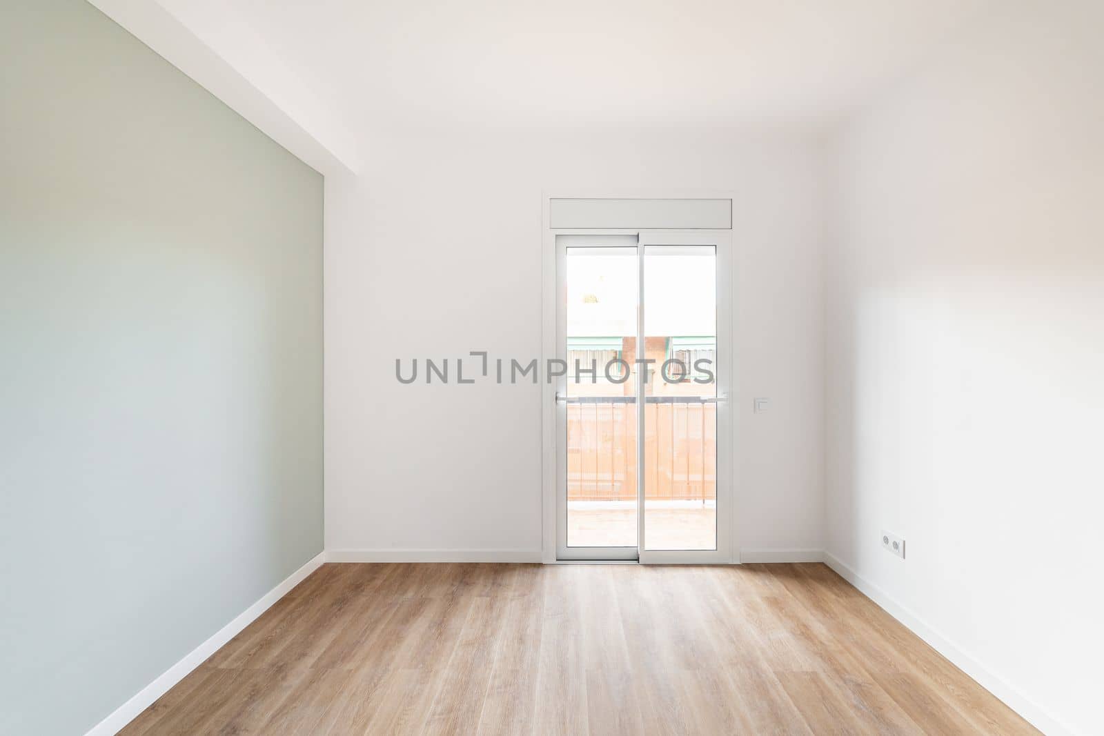 An empty bright room for living with access to balcony and view of neighboring house. Room has transparent glass door with access to balcony. Gray wall and wooden laminate floor create cozy feeling