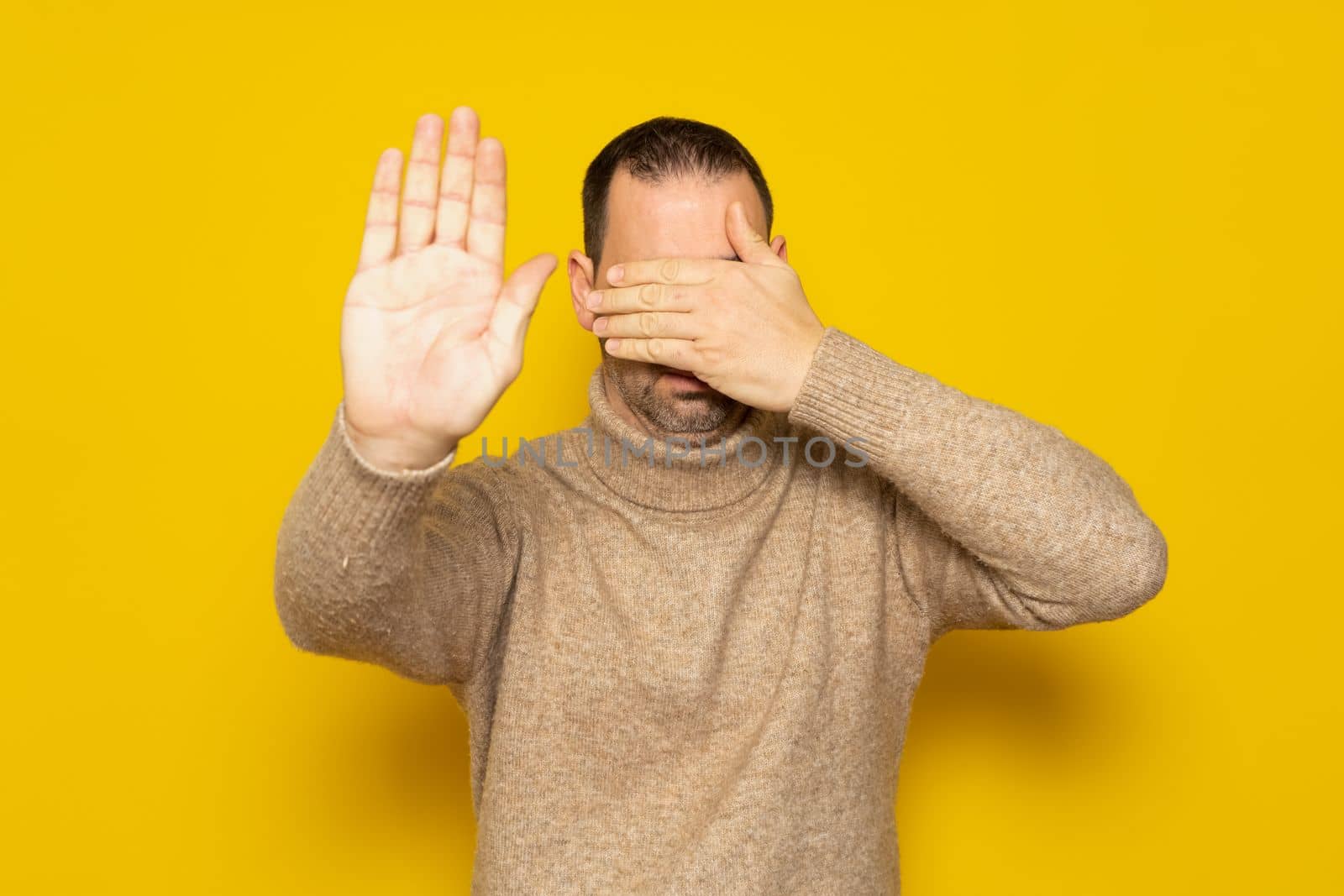 Man in his 40s with a beard wearing a turtleneck covering his eyes with his hands and making a stop gesture with an expression of sadness and fear. Embarrassed and negative concept.