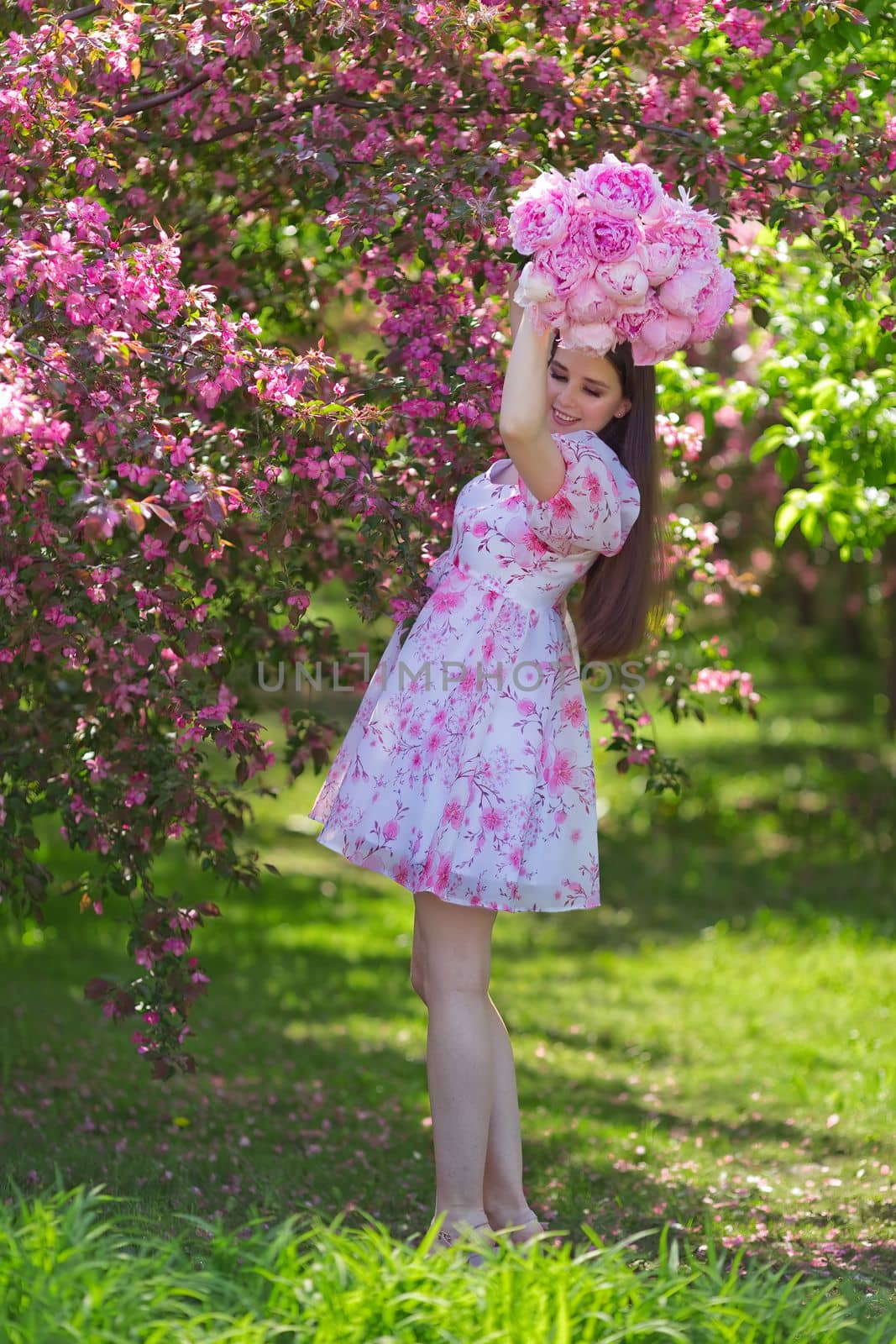 Happy girl with long hair, brunette, in a light pink dress, holding a bouquet of large pink peonies over her head, standing near pink blossom apple trees, in the garden on a sunny day. Space for copying. Vertical