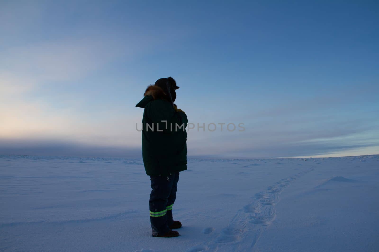Man in winter clothing standing on snow watching the weather on tundra landscape while holding a rifle, near Arviat, Nunavut, Canada