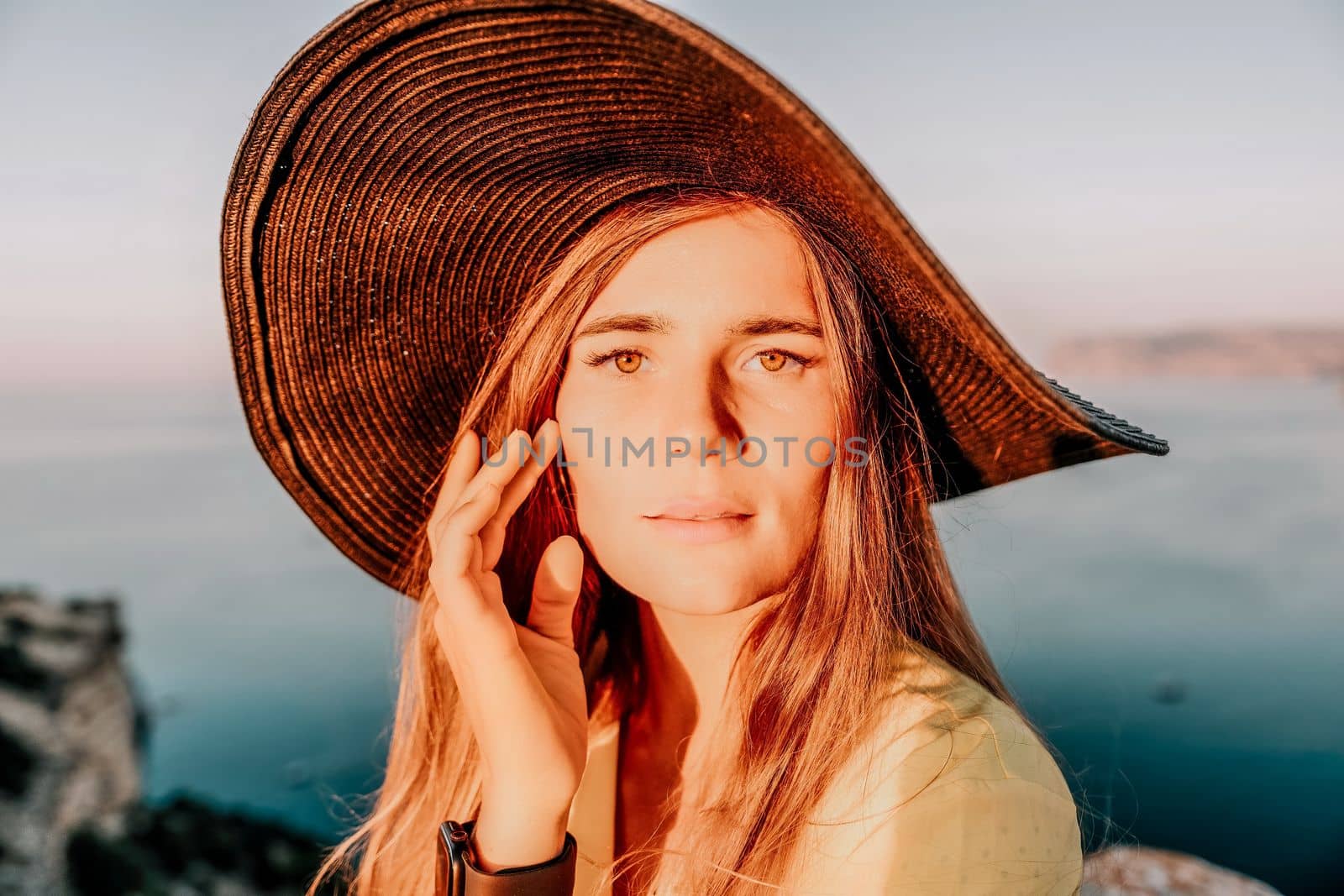 Portrait of happy young woman wearing summer black hat with large brim at beach on sunset. Closeup face of attractive girl with black straw hat. Happy young woman smiling and looking at camera at sea