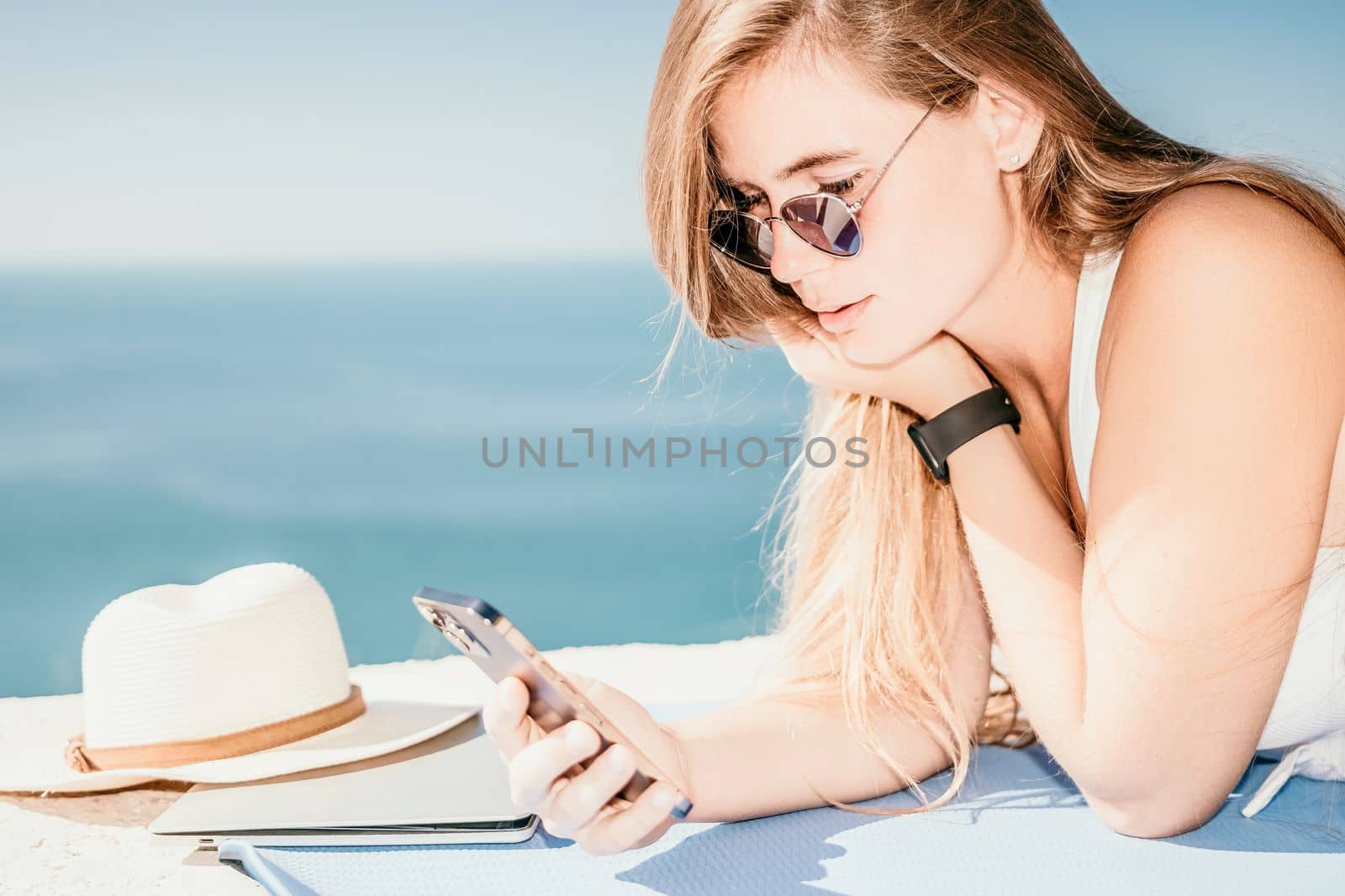 Successful business woman in yellow hat working on laptop by the sea. Pretty lady typing on computer at summer day outdoors. Freelance, travel and holidays concept.