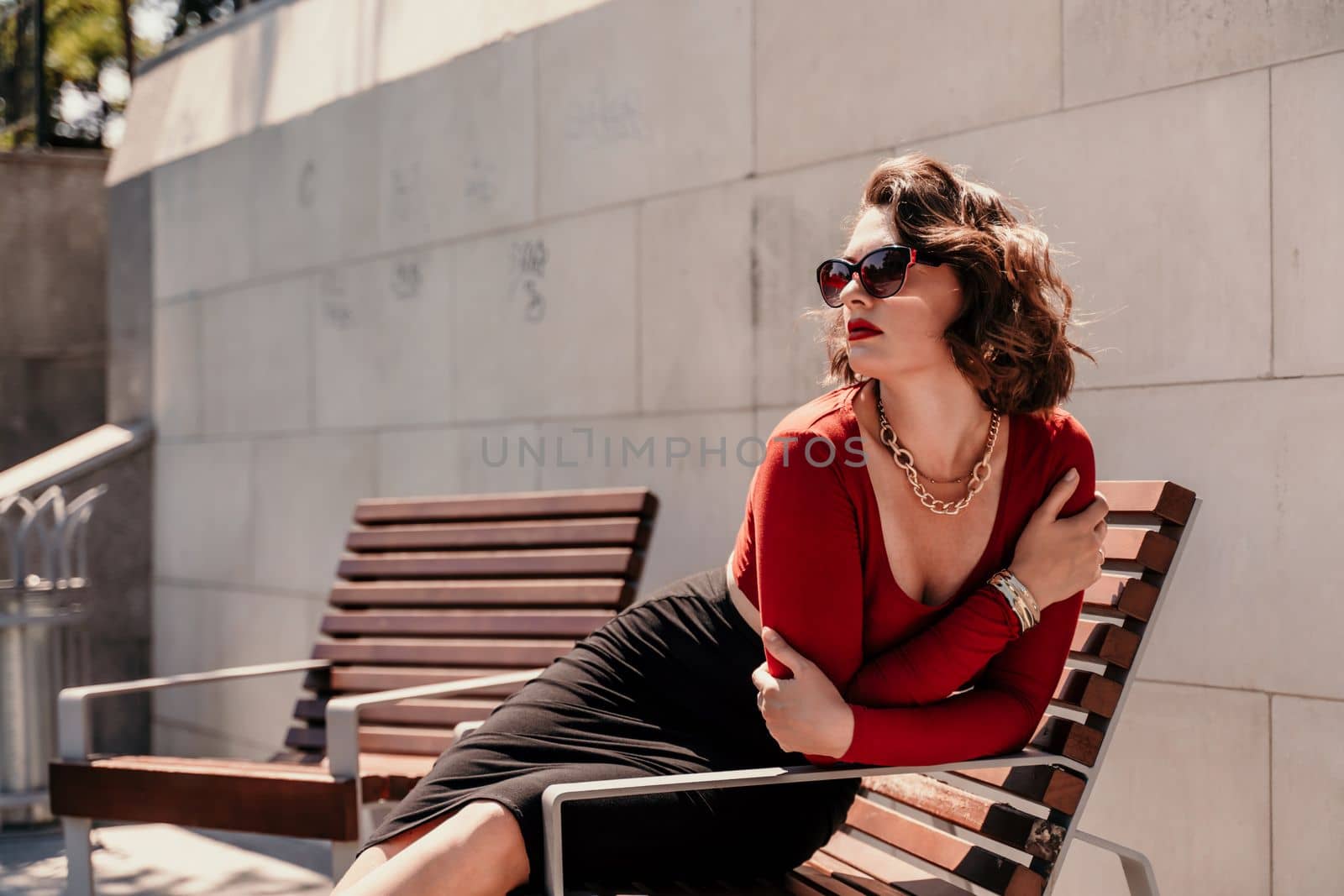 Portrait of a woman on the street. An attractive woman in glasses, a red blouse and a black skirt is sitting on a bench outside. by Matiunina
