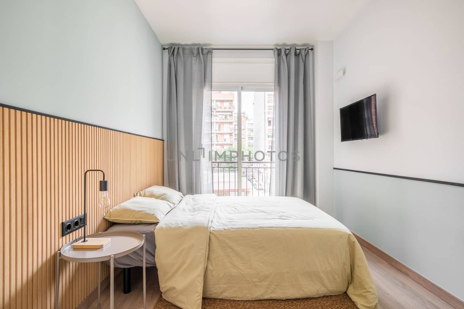 Bedroom with large bed for two persons against backdrop of glass sliding door with access to small balcony. Curtains on door protect from bright sun during day. On wall is TV for watching movies