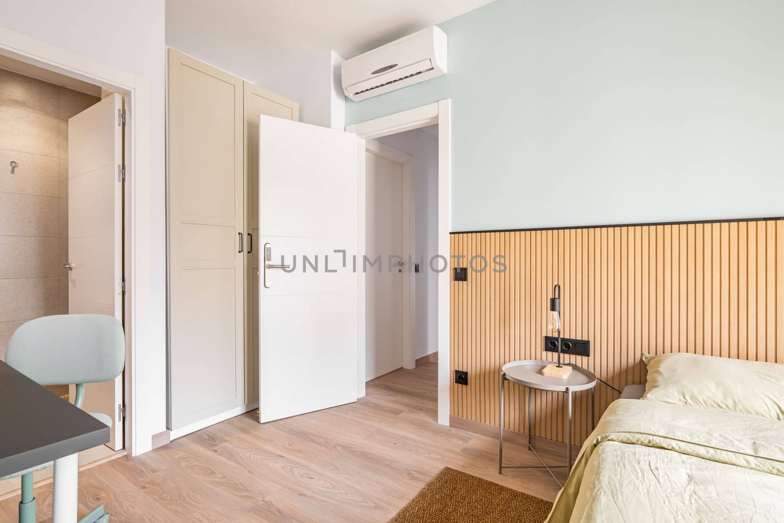 Bedroom with bed, wooden wardrobe built into wall, table and chair for work. Air conditioning on door for comfortable temperature in room. Near bed there is a table with night lamp. by apavlin