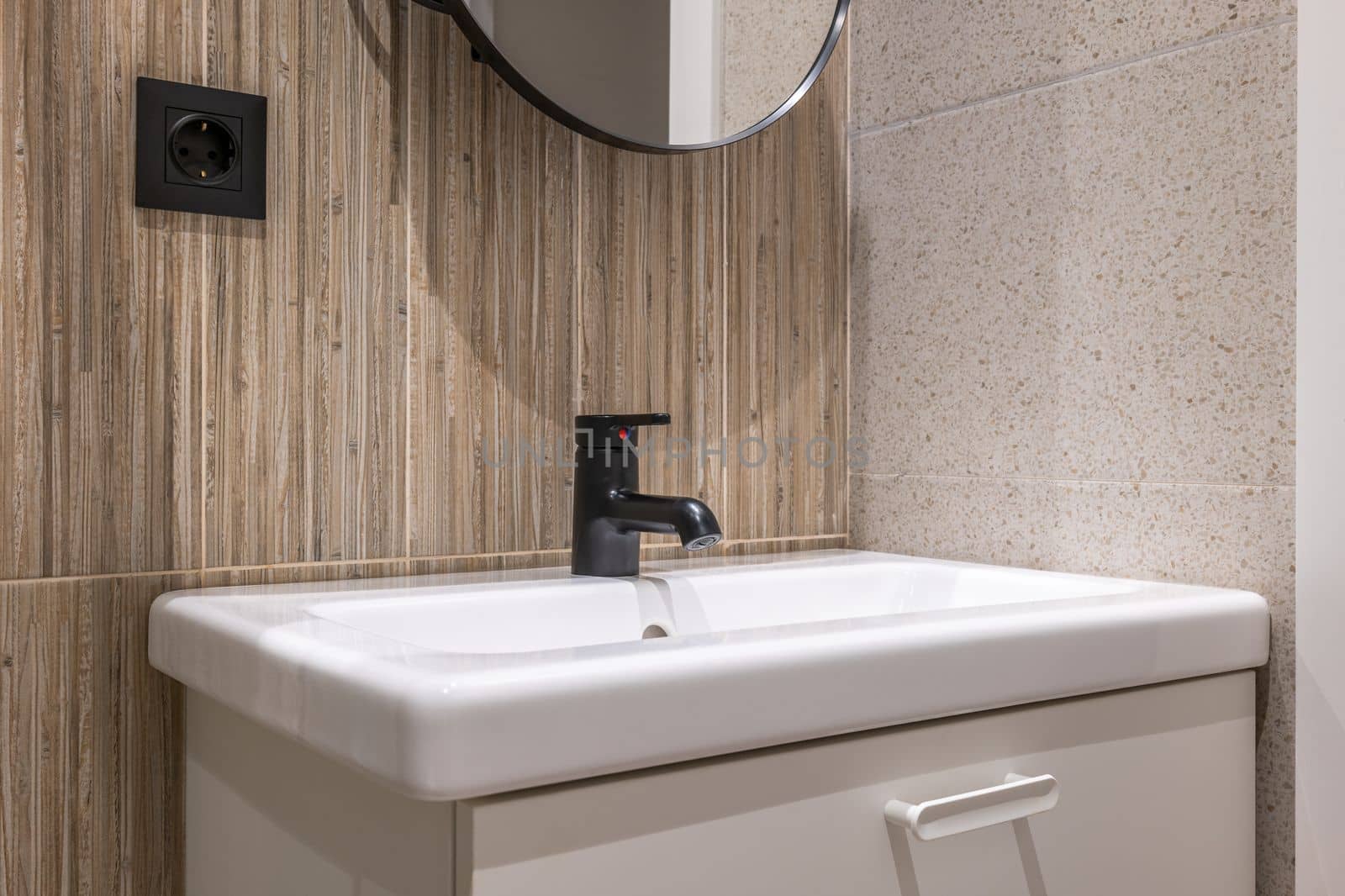 Bathroom sink on vanity is illuminated by bright light from ceiling. Walls are combination of gray granite and wood grain marble tiles. Above sink is mirror in black frame, like faucet and an outlet. by apavlin