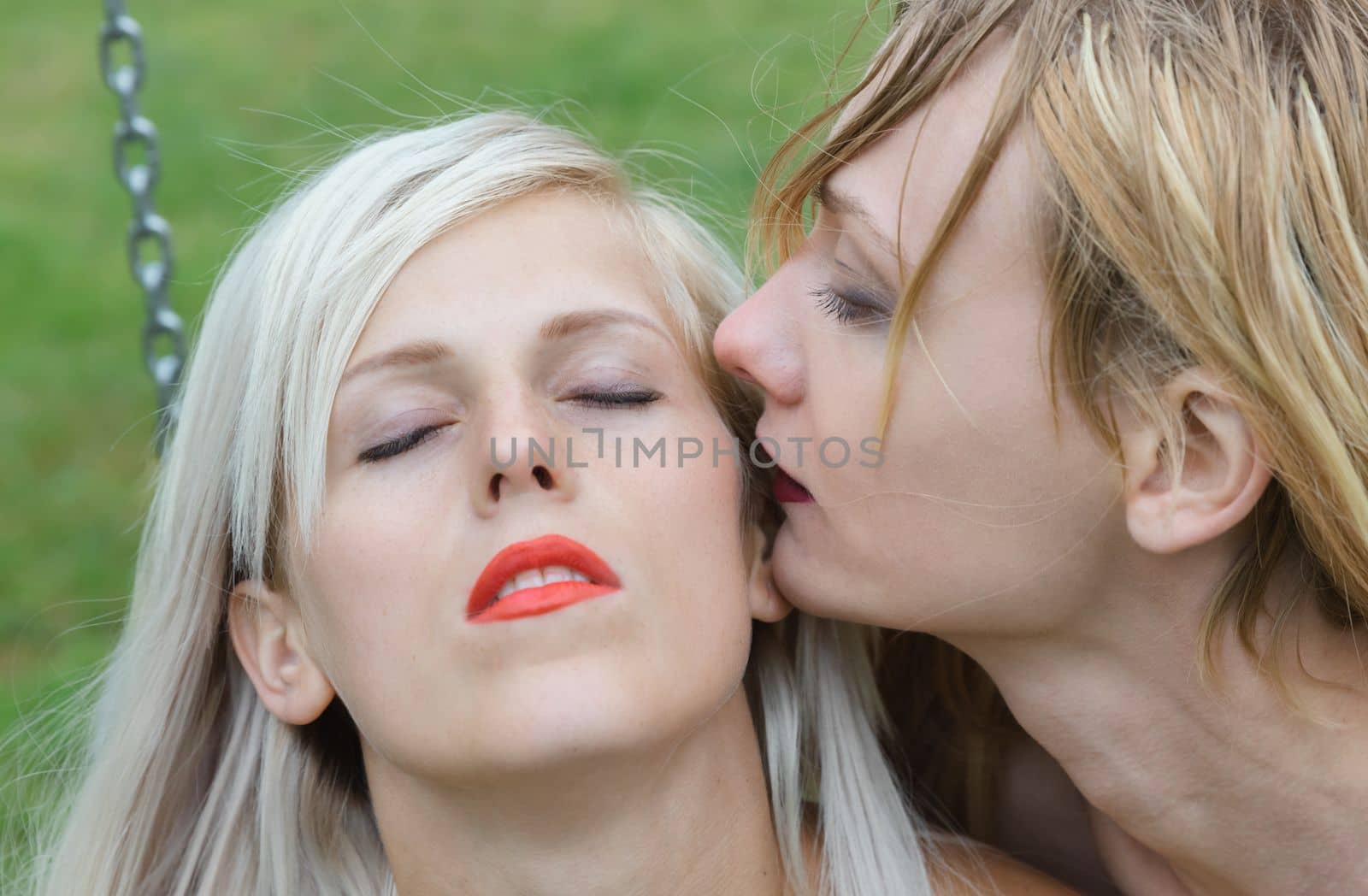 Two women, one blonde and one brunette, are  sharing a romantic moment outdoors on a sunny day. They express their love and passion for each other with a kiss, conveying a beautiful and tender emotion