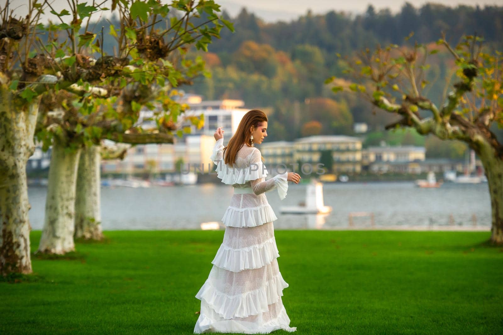 A bride in a white wedding dress in a park in an Austrian town with large trees at sunset by Lobachad