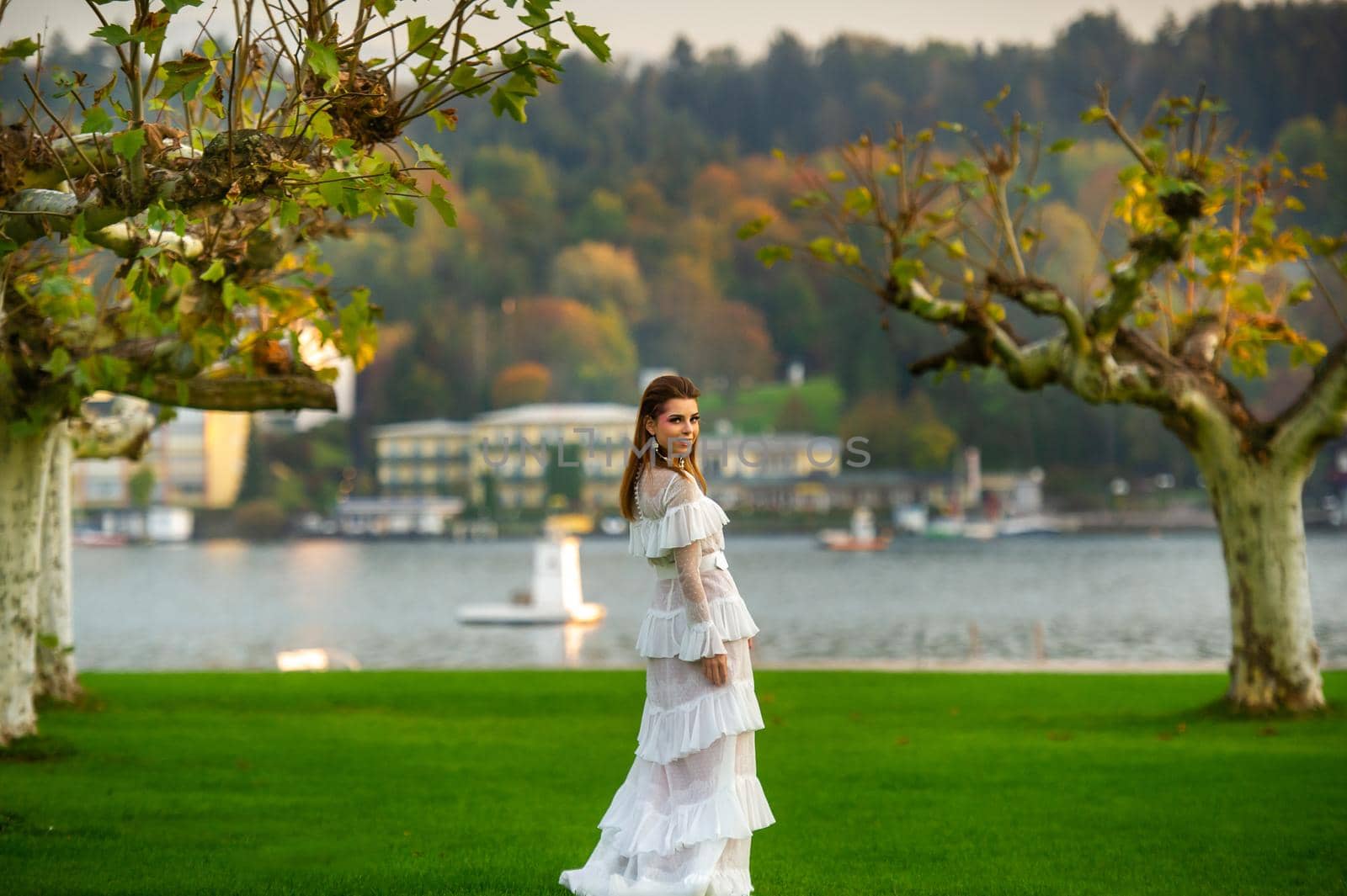 A bride in a white wedding dress in a park in an Austrian town with large trees at sunset by Lobachad