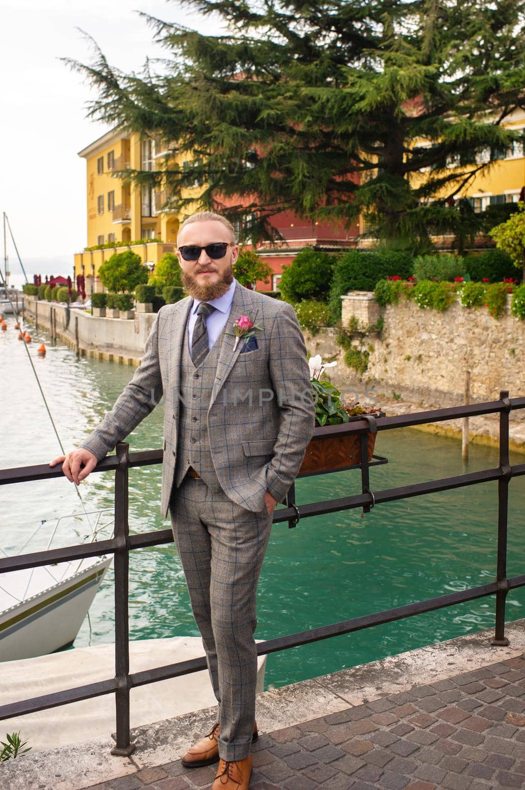 A man with a beard in a strict grey three-piece suit with a tie in the old town of Sirmione, a Stylish man in a grey suit in Italy by Lobachad