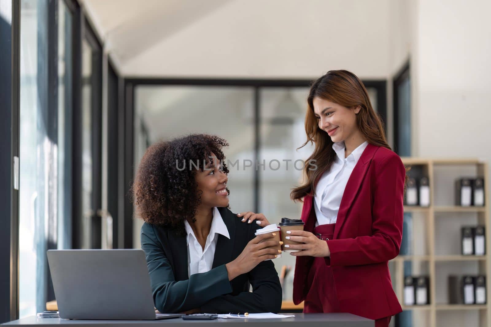 Two Asian business women enjoy chatting during coffee breaks at their desks in a comfortable conference room by nateemee