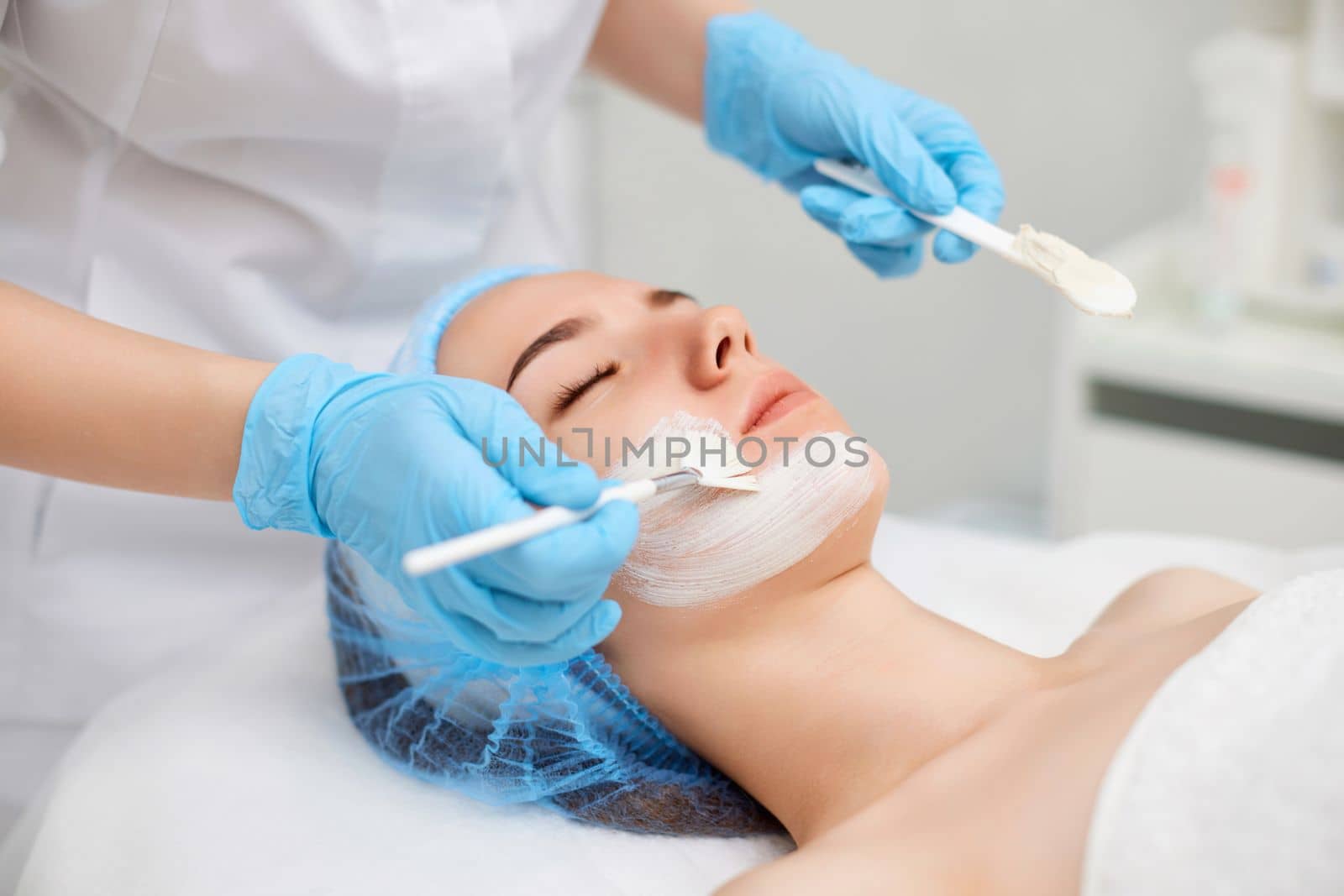 beautician applies the mask to the face of beautiful young woman in the spa salon. Anti-aging, facial skin care concept.